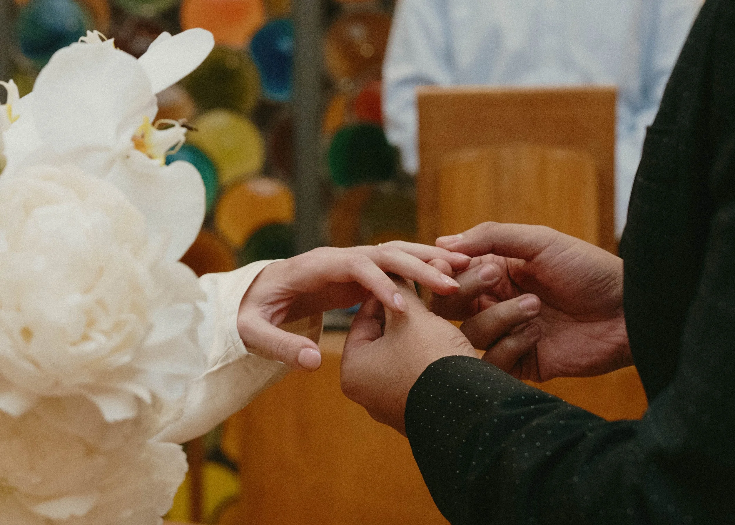 Close-up of two people holding hands, one wearing a white shirt and the other wearing a black shirt, in a setting with colorful objects in the background.