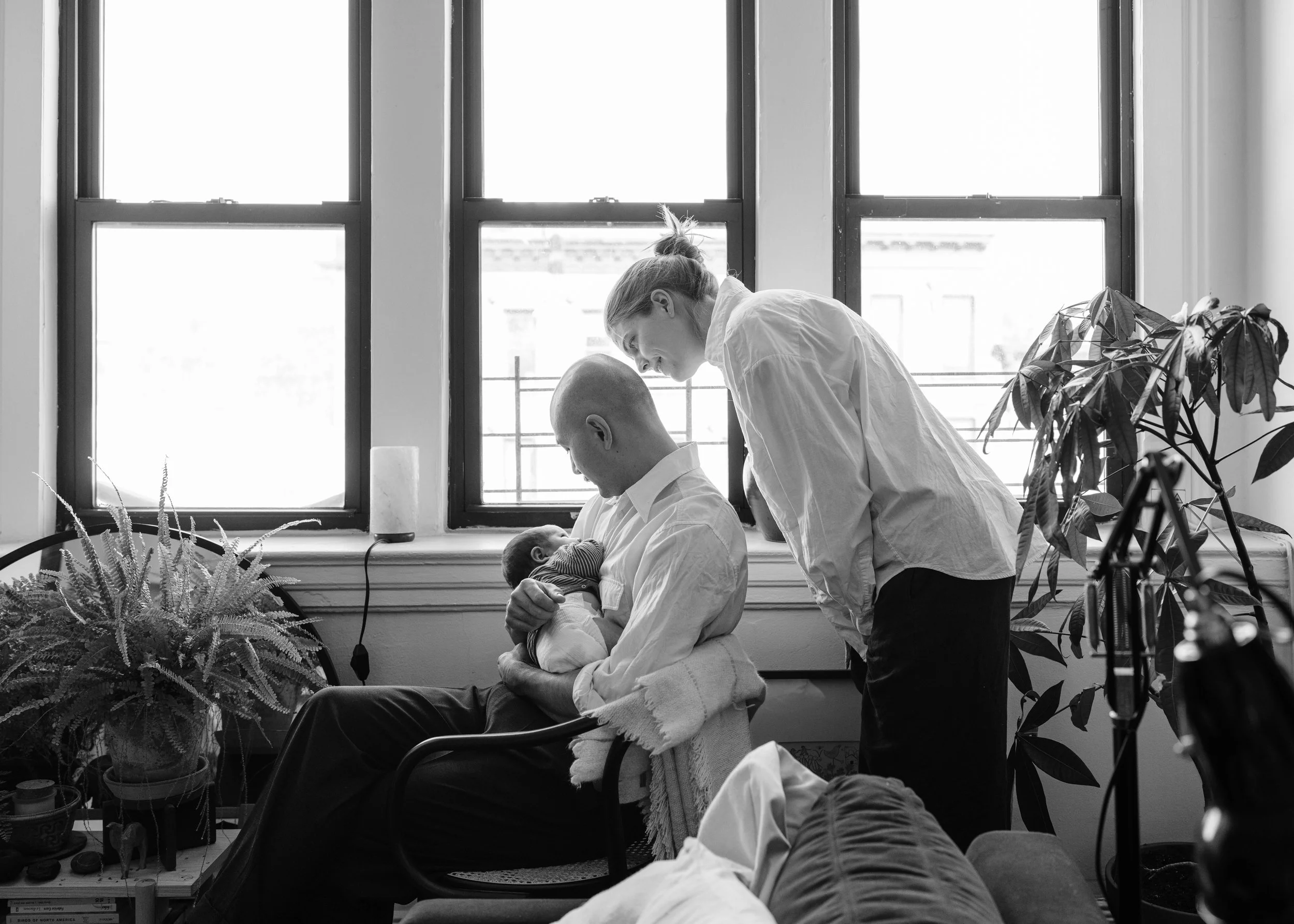 Man holding a newborn baby on his lap, woman leaning over, all in a room with large windows and houseplants.