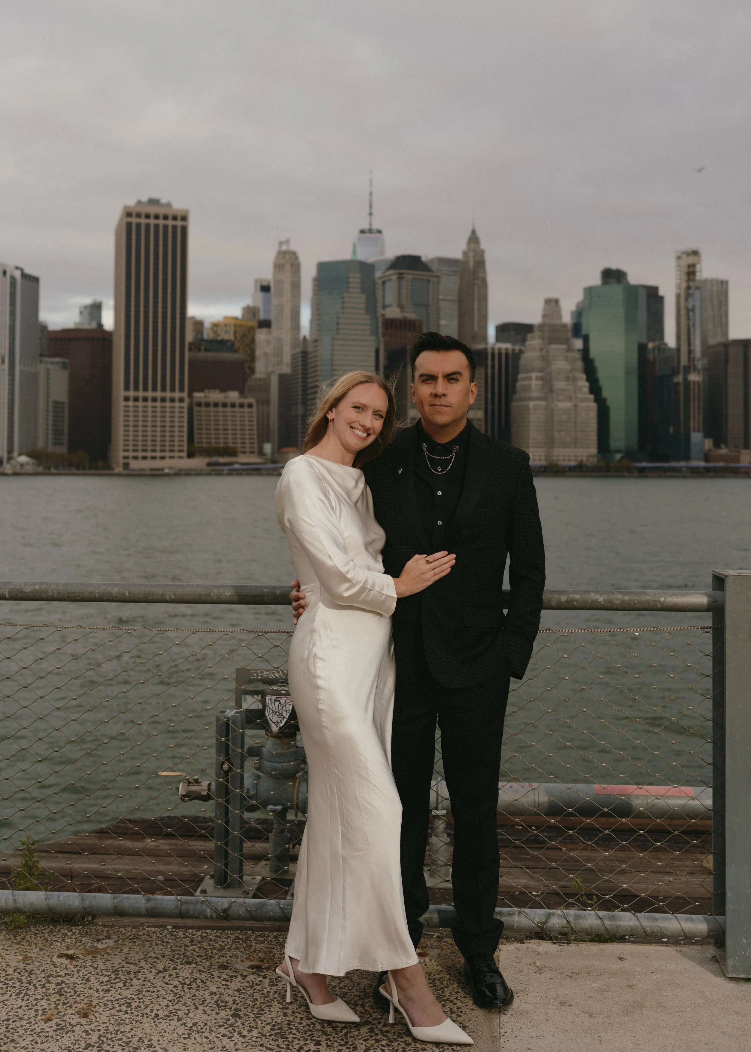 A woman and a man standing together outdoors near the water with a city skyline in the background. The woman is wearing a white satin dress and high heels, smiling, with her arm around the man's shoulder. The man is dressed in a black suit and black 