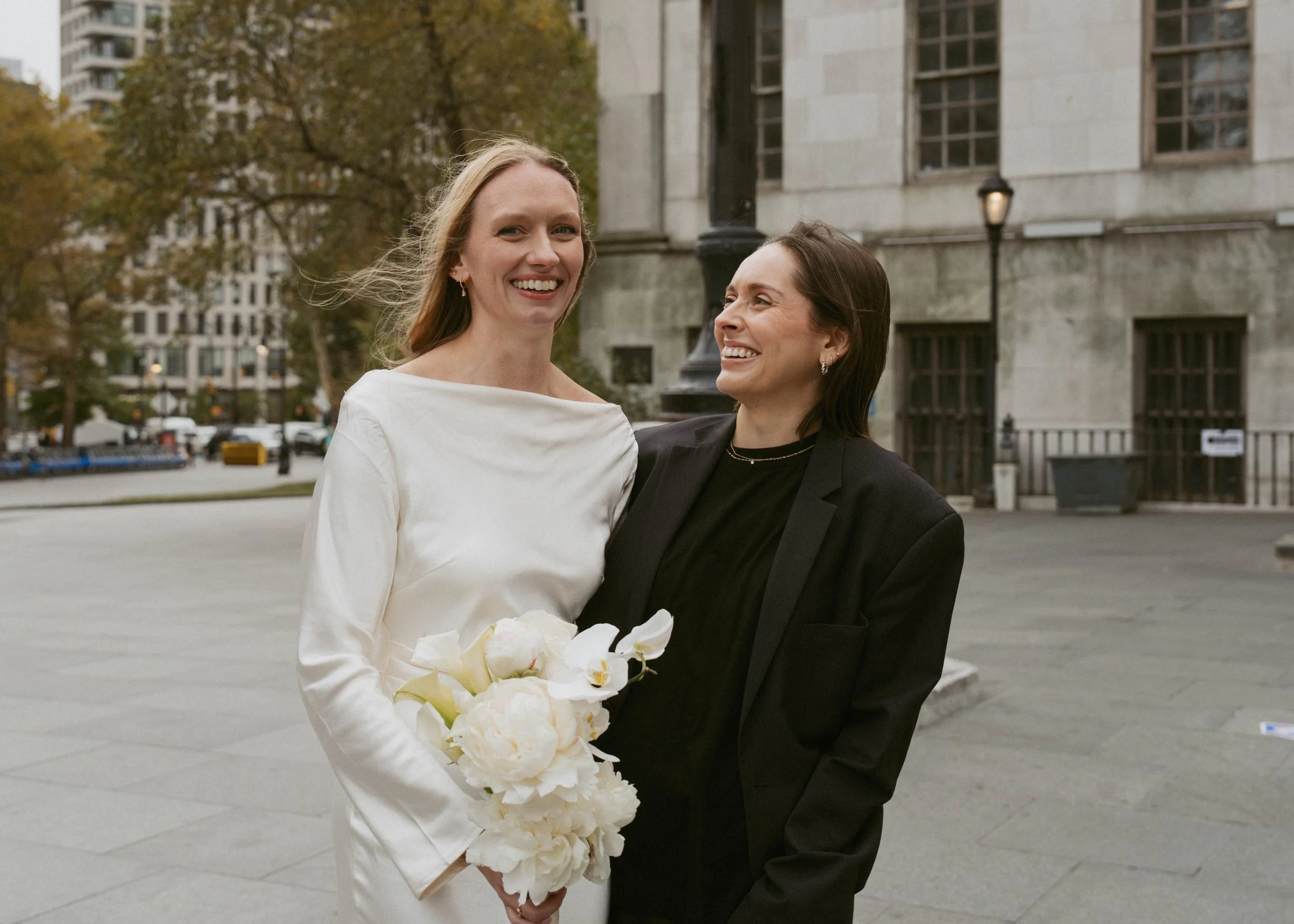 Two women smiling and standing close together outside in an urban area; one is holding a bouquet of white flowers, and the background shows trees, buildings, and street lamps.