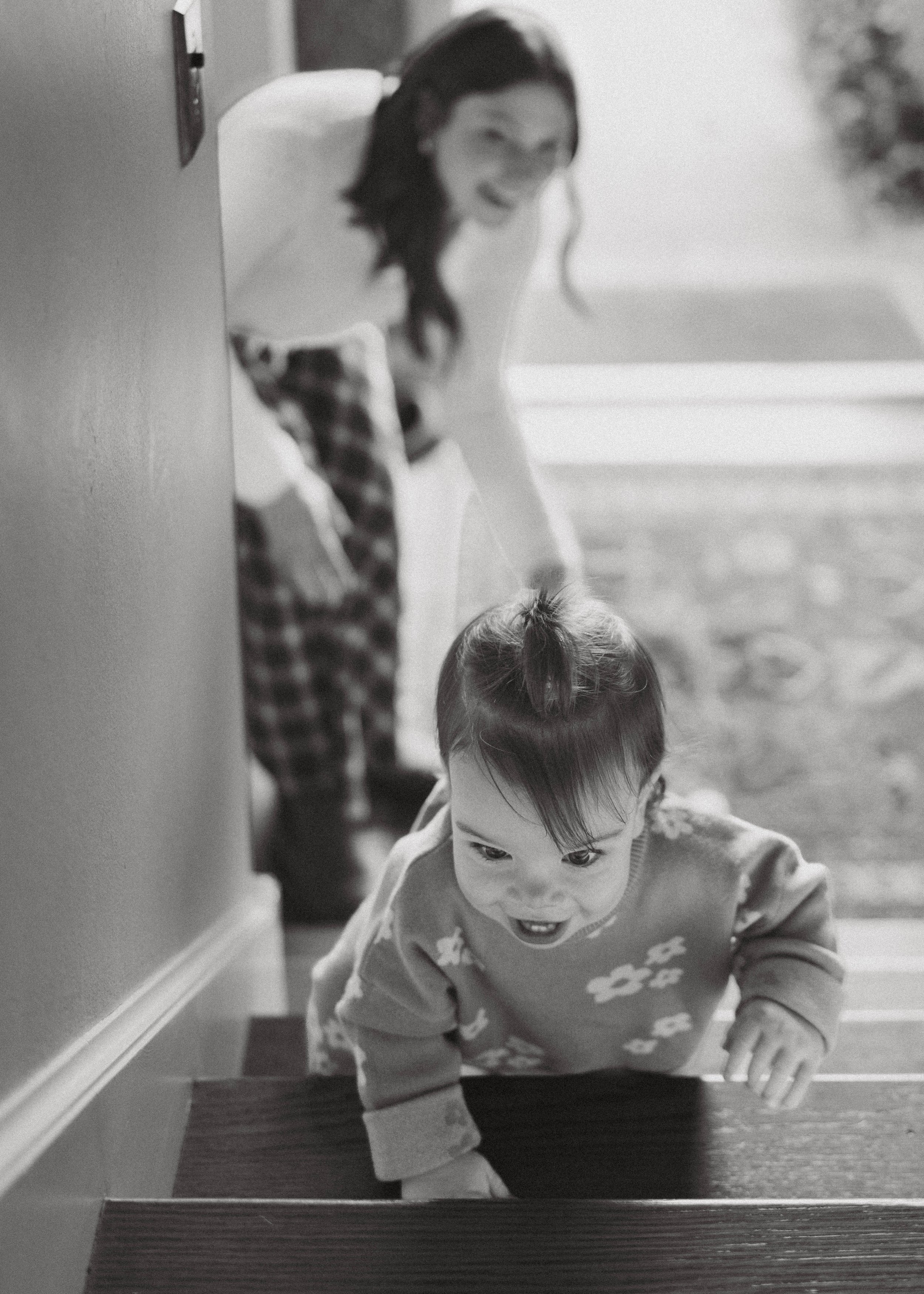 Black and white photo of a young boy climbing up a set of stairs, with a woman smiling behind him, visible in the background.