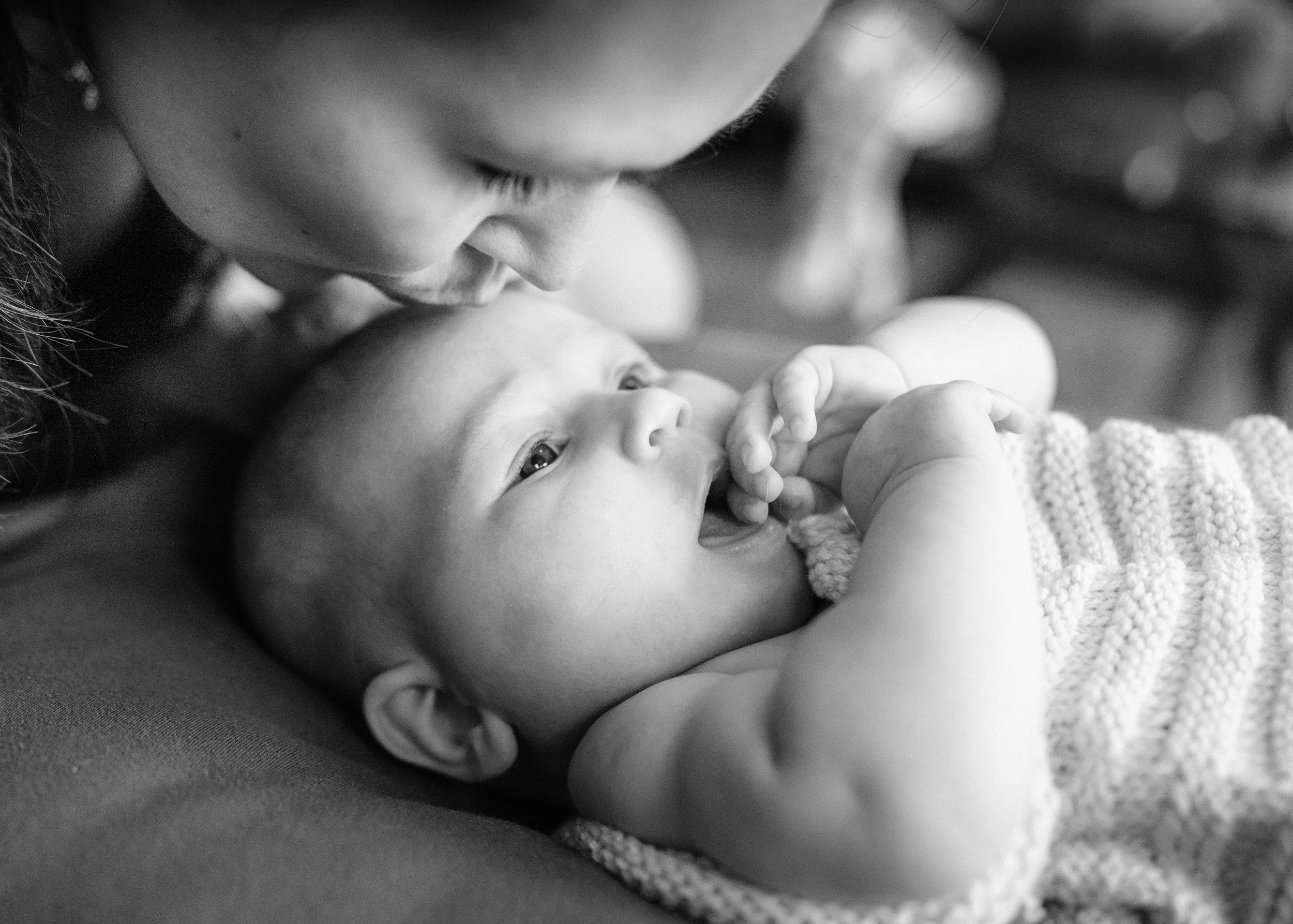 A black and white photo of a woman close to a baby, holding the baby and touching their face, with the baby lying on a soft surface, looking up and sucking on fingers.