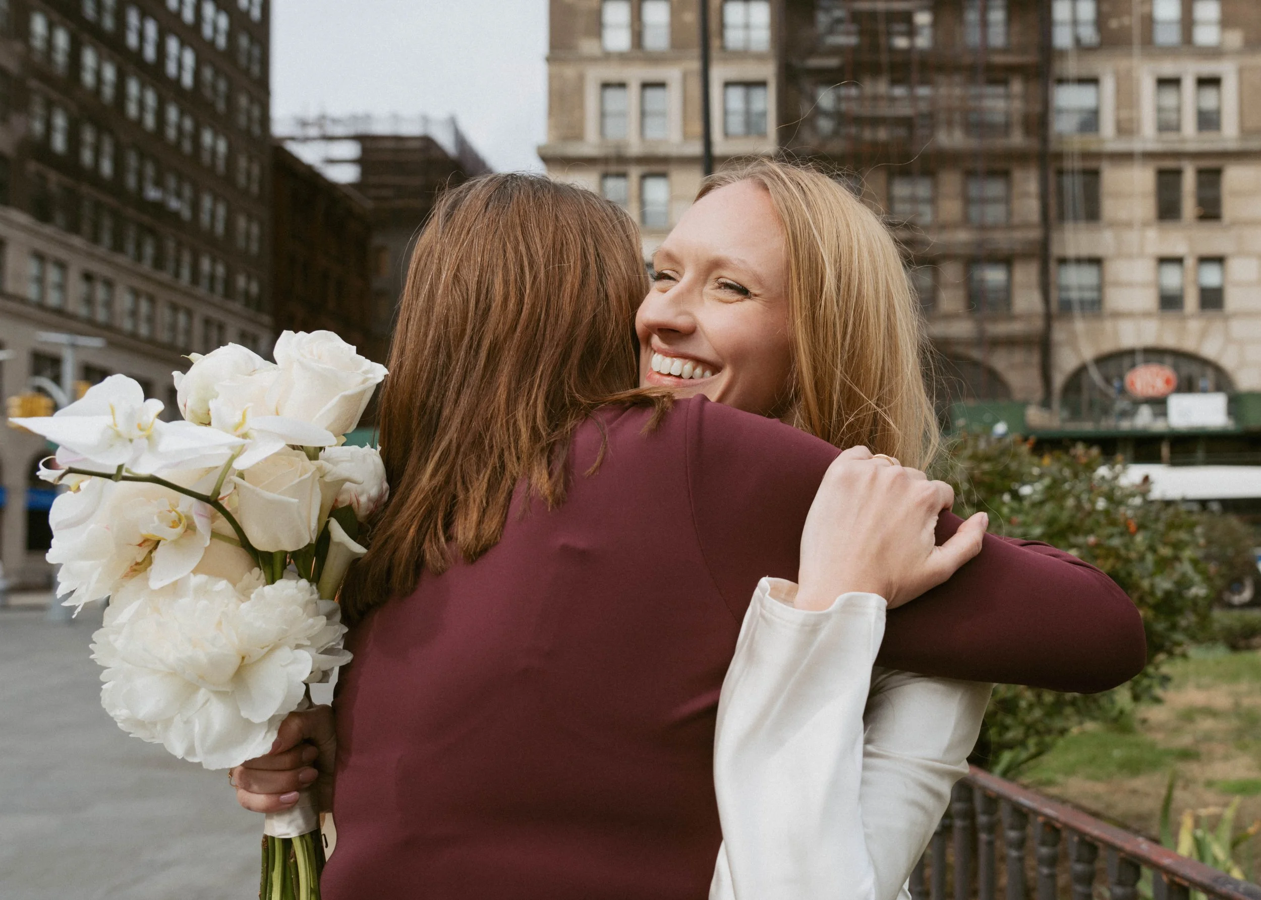 Two women hugging on a city street, one woman holding a bouquet of white flowers, smiling and embracing warmly.