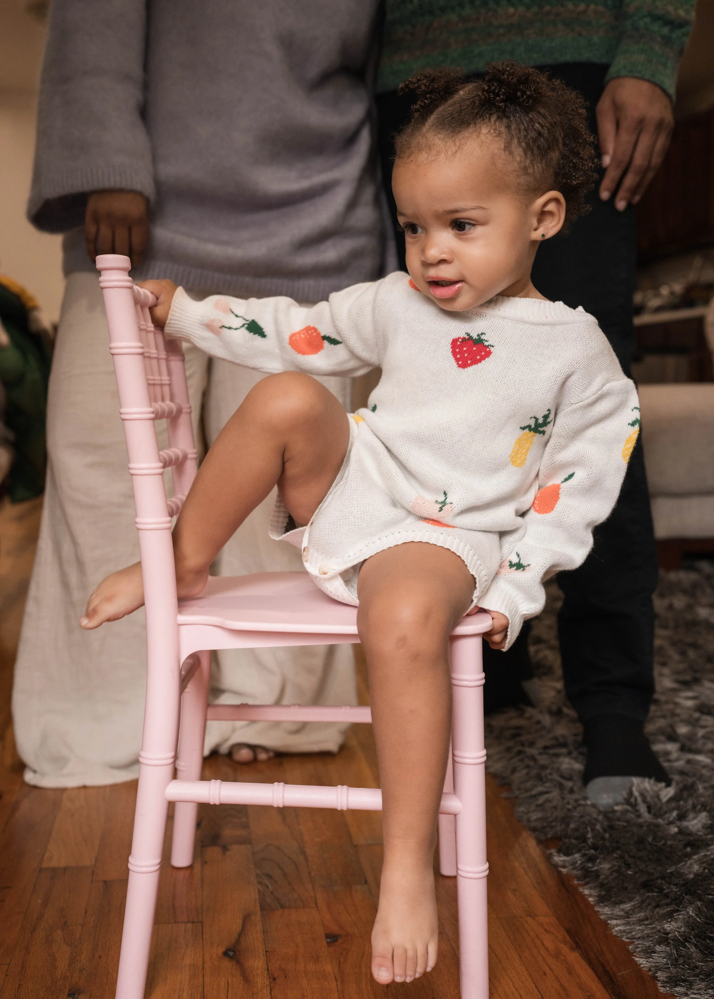 A young girl with curly hair sitting on a pink wooden chair indoors, wearing a sweater with colorful fruit prints, with adults standing behind her.