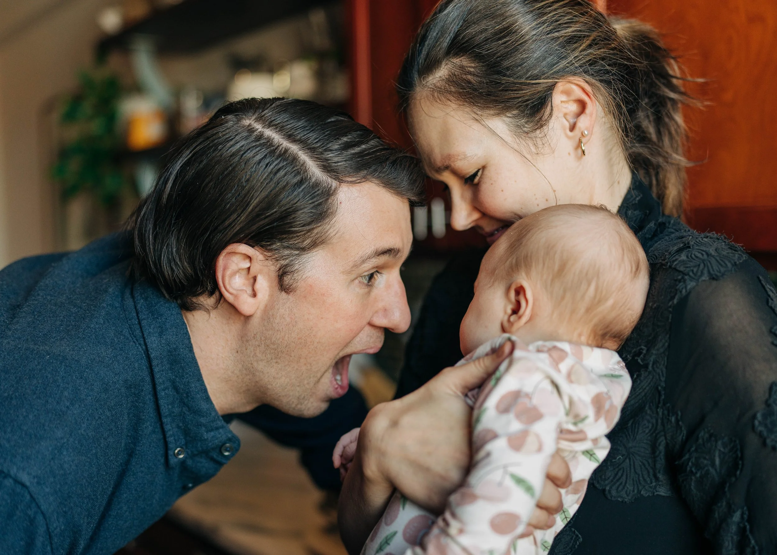 A man, woman, and baby sharing a close, joyful moment indoors. The man and woman are touching foreheads, smiling, and looking at the baby, who is being held and cuddled by the woman.