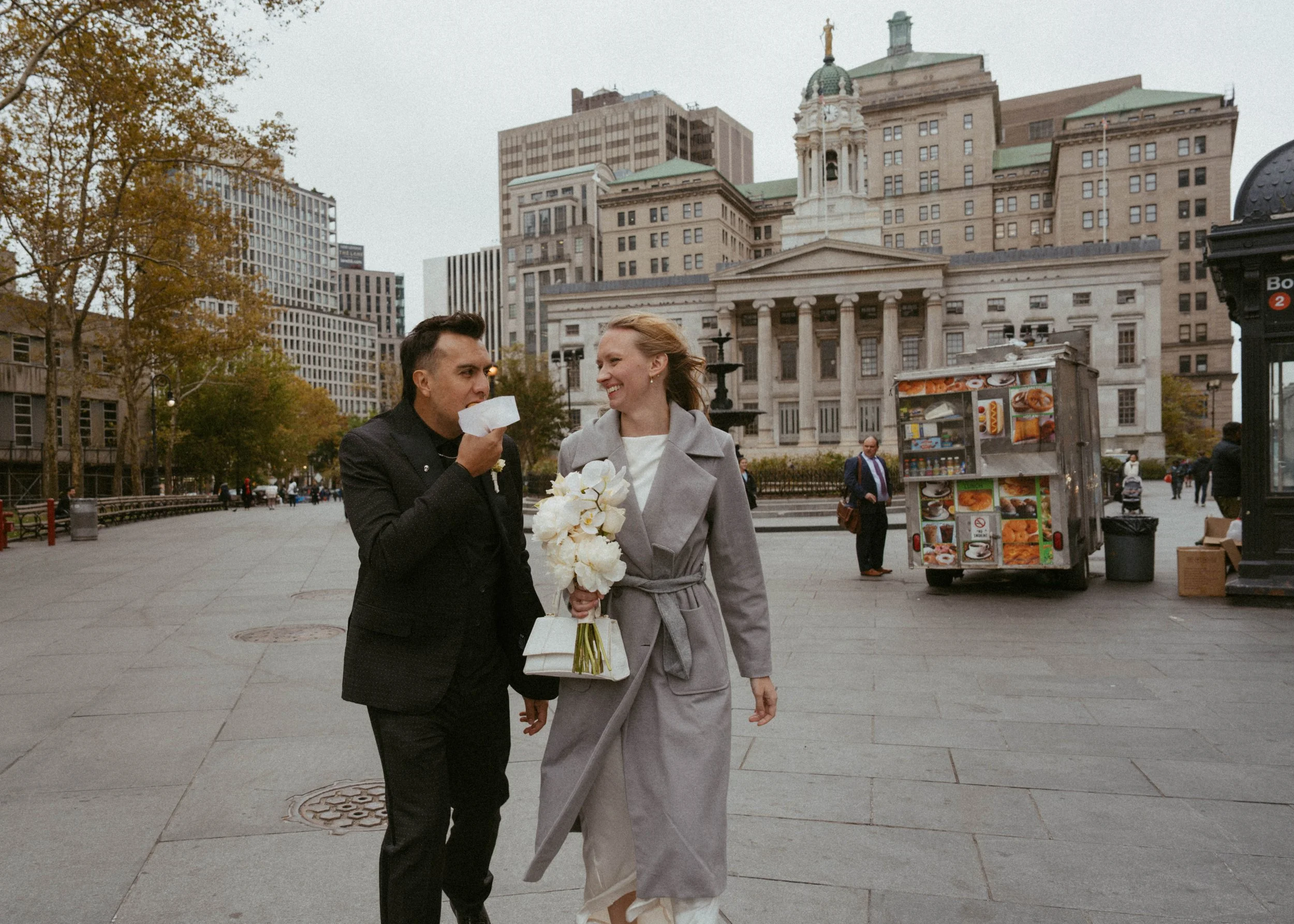 A man and woman walking together in an urban park, with the man licking a white paper, and the woman smiling while holding a bouquet of white flowers, in front of a historic building and city skyline.