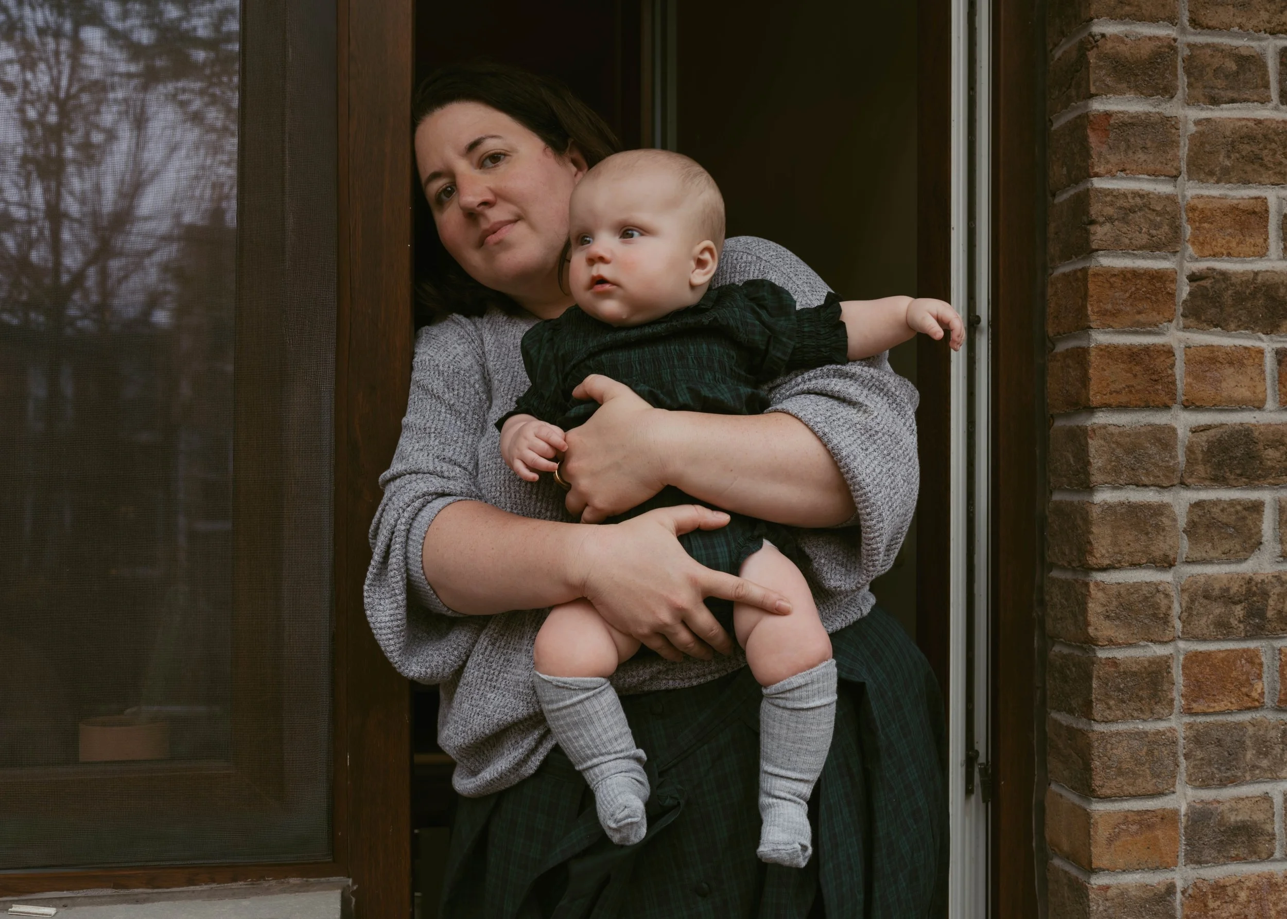 A woman holding a young child at a doorway with brick and wood framing, outside a house.