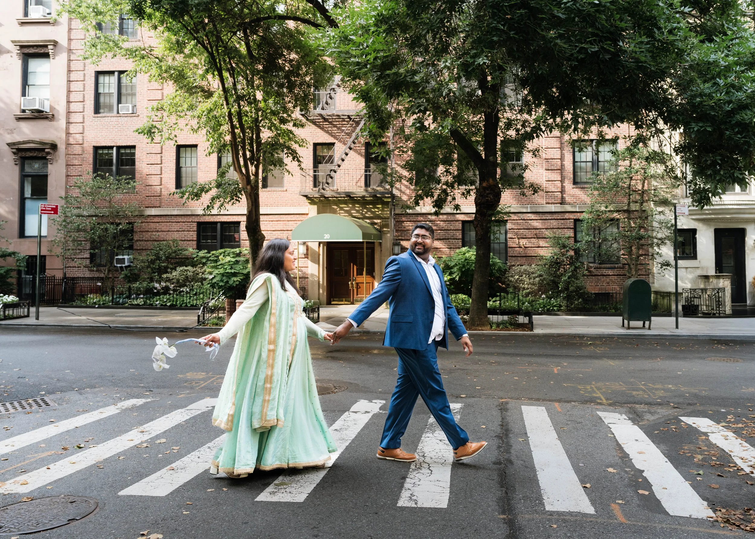 A man and a woman holding hands, walking across a crosswalk on a city street. The woman is wearing a light green traditional dress with gold accents, and the man is wearing a blue suit with brown shoes. They are smiling and looking at each other as they walk. There are trees and brick apartment buildings in the background.
