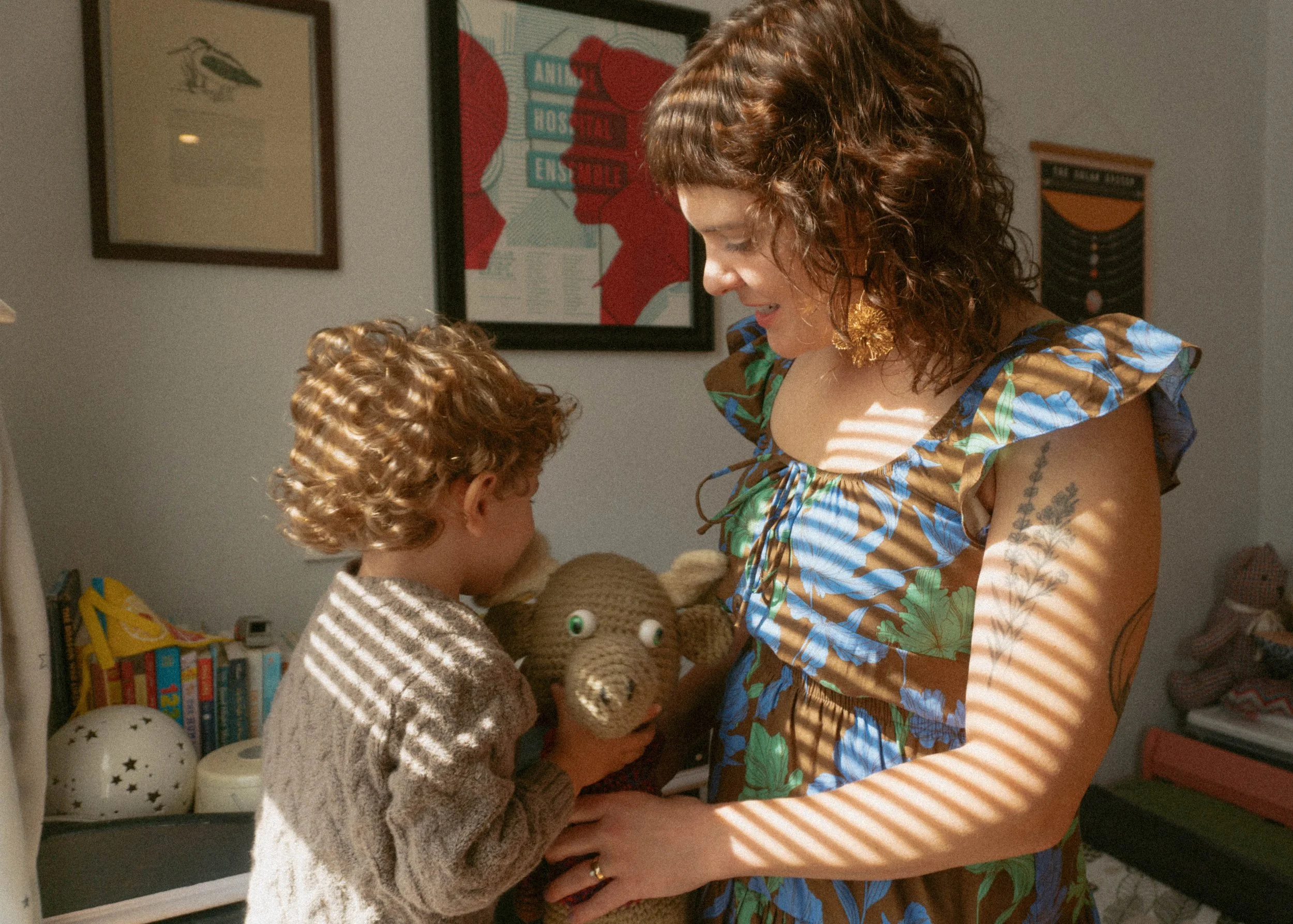 A woman and a young boy with curly hair are playing with a crocheted stuffed animal. The woman, wearing a colorful, patterned dress, is smiling while looking at the boy. The boy is in a beige sweater and is holding the stuffed animal close to his fac