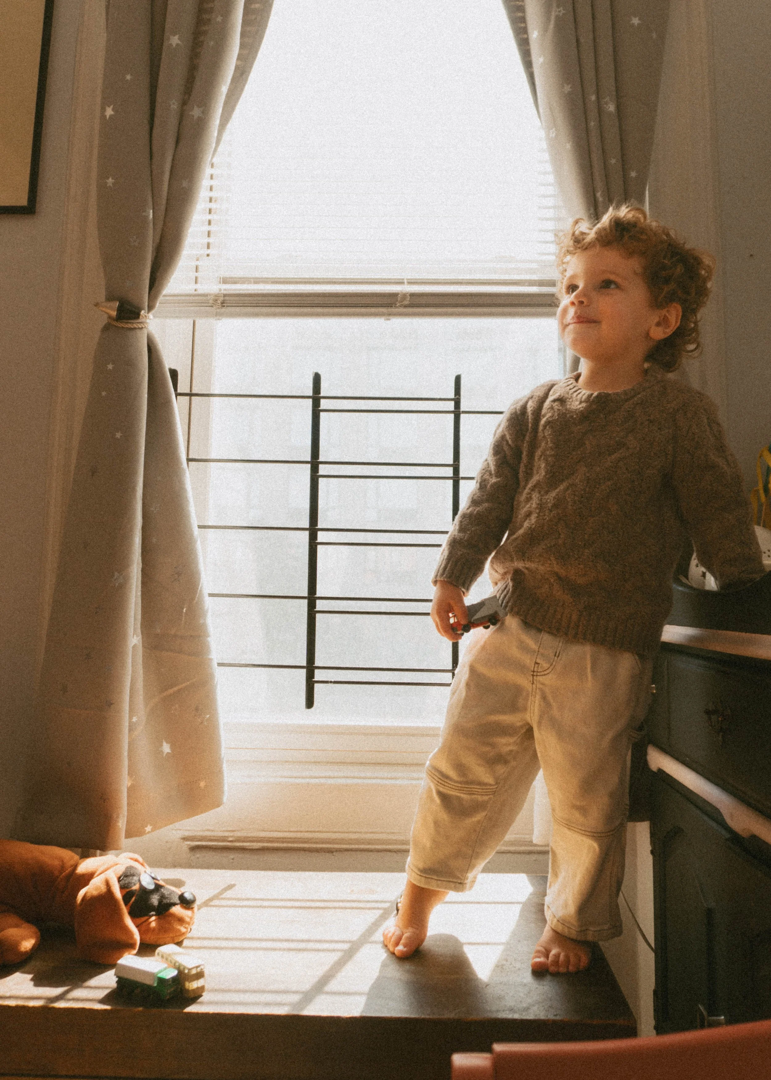 A young boy with curly hair, wearing a brown sweater and beige pants, standing on a kitchen counter near a window with beige curtains, holding a toy car in his right hand, looking thoughtful.