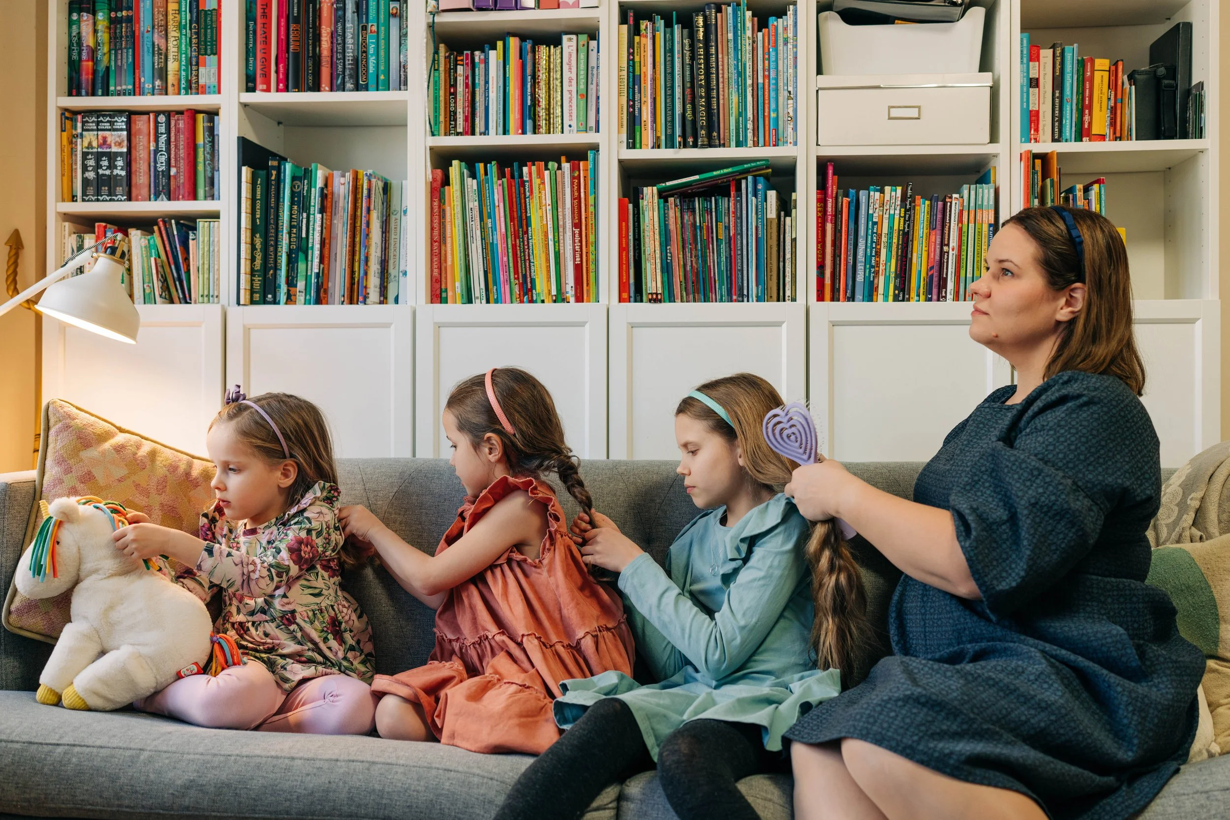 A woman and three young girls sitting on a couch, playing with a plush unicorn toy, with a bookshelf filled with books in the background.