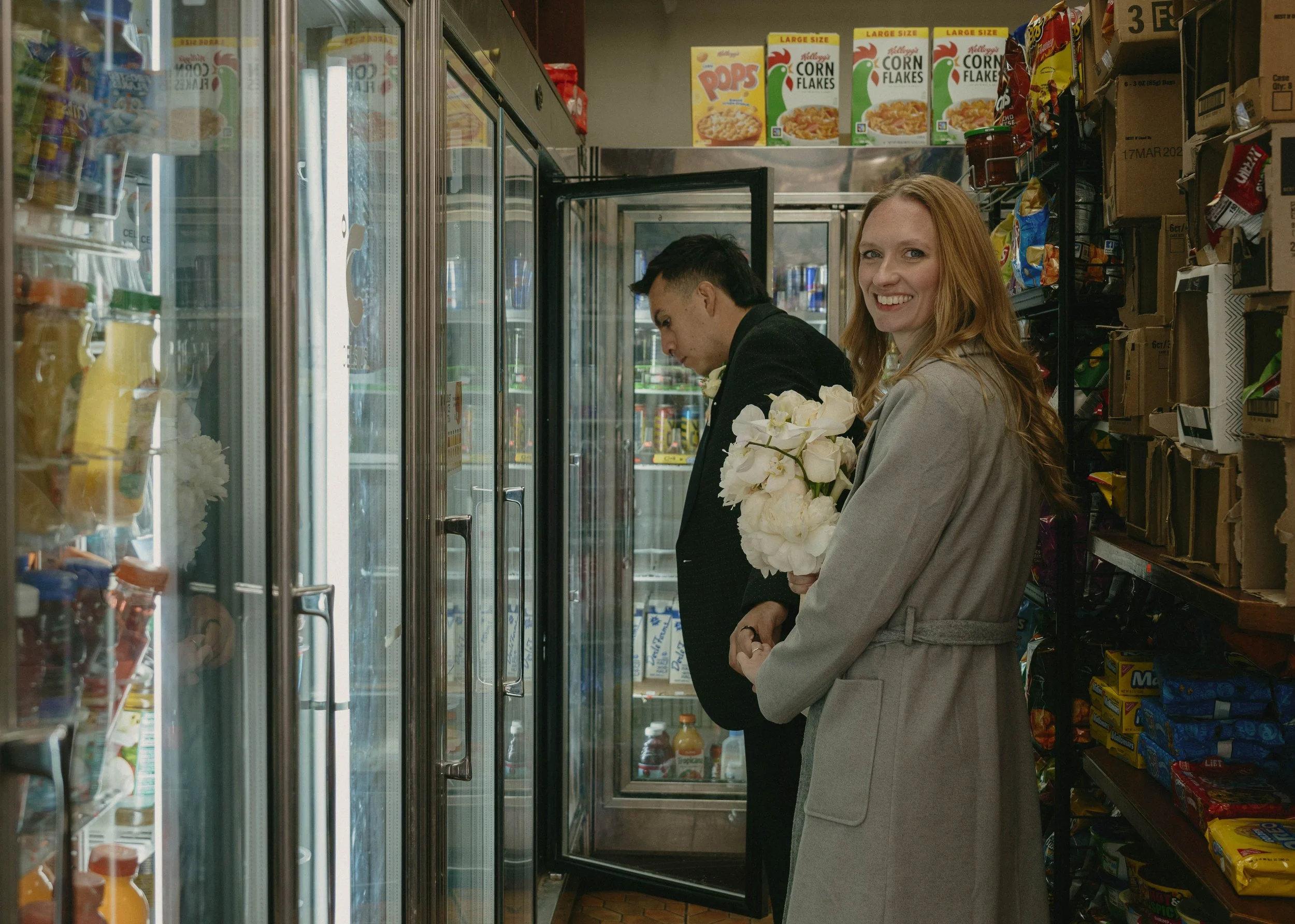 A woman in a gray coat holding white flowers smiling at the camera inside a grocery store aisle, with a man in a black jacket looking down at the fridge door behind her.