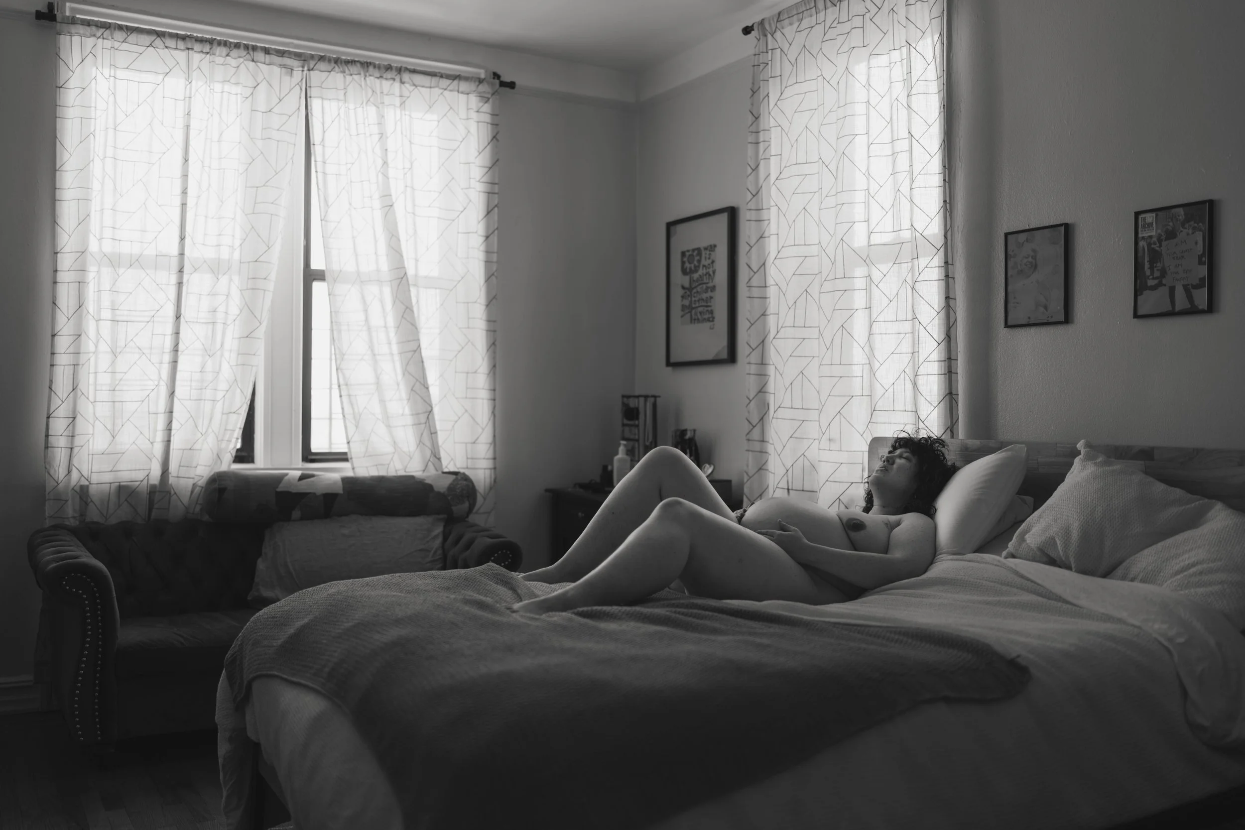A woman lying on a bed in a bedroom, looking up, with natural light coming through windows with patterned curtains.