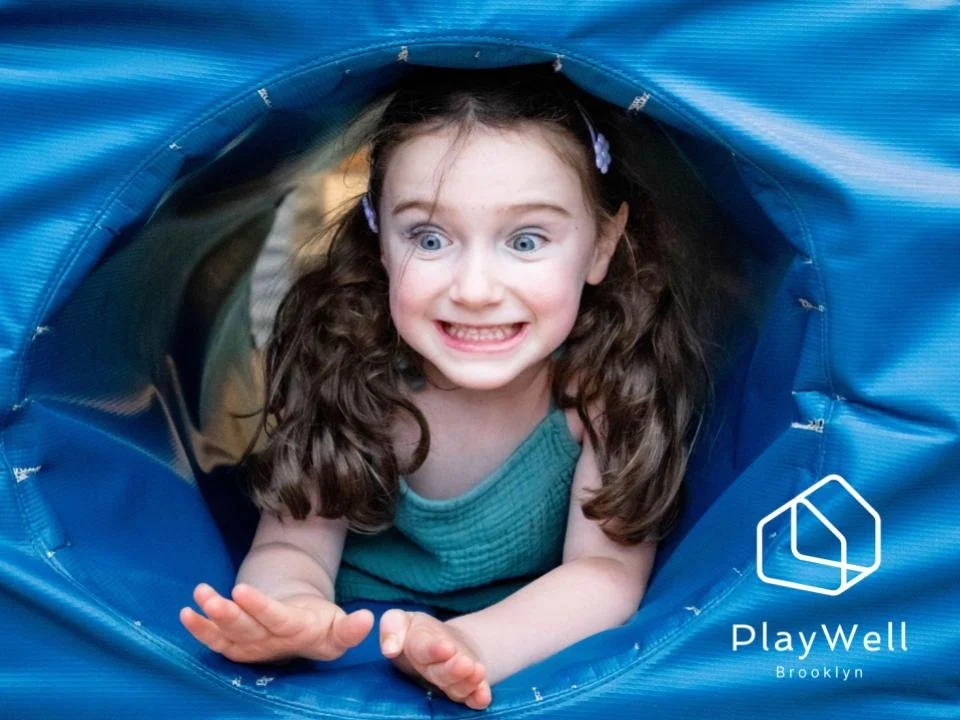 A young girl with curly brown hair and blue eyes smiling while crawling through a blue tunnel.