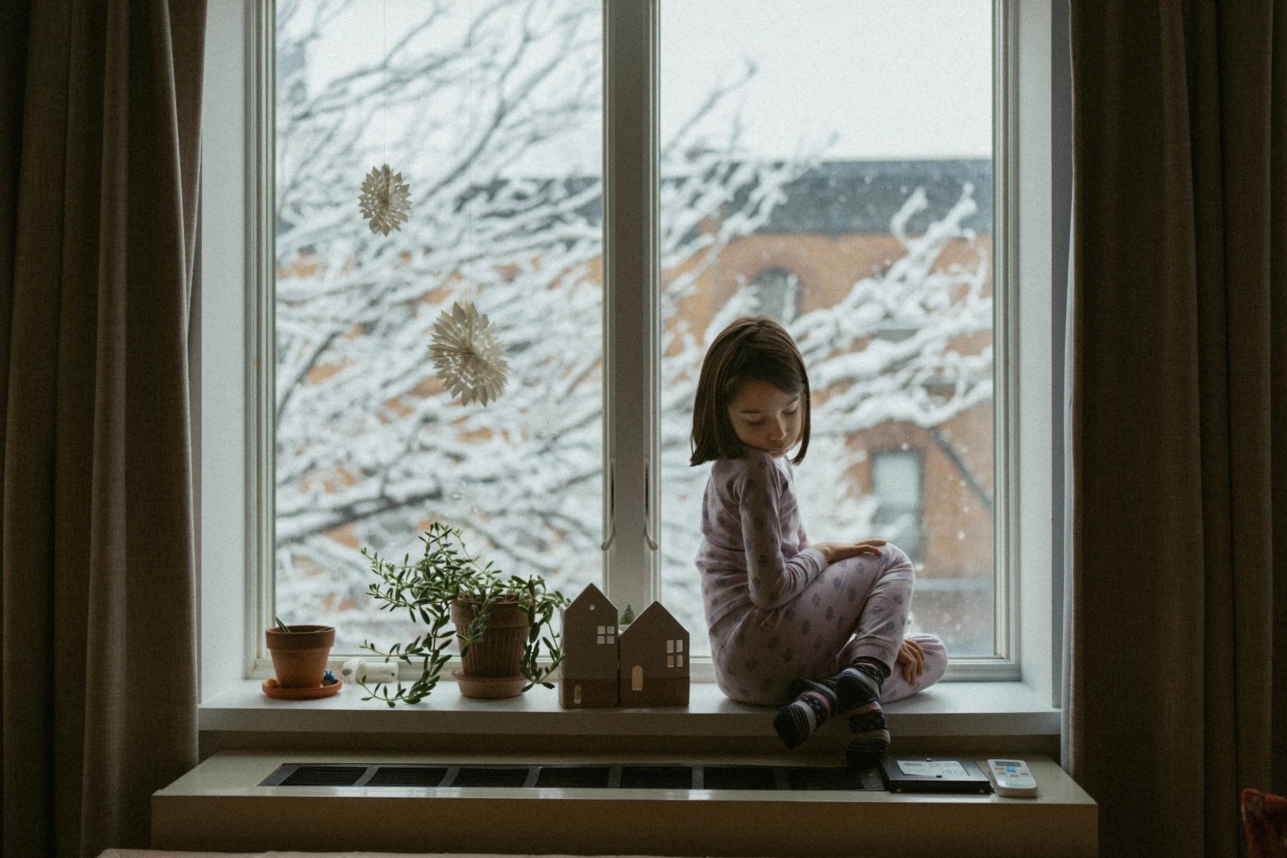 A young girl sitting on a windowsill in pajamas, looking down with snow-covered trees and houses visible outside.