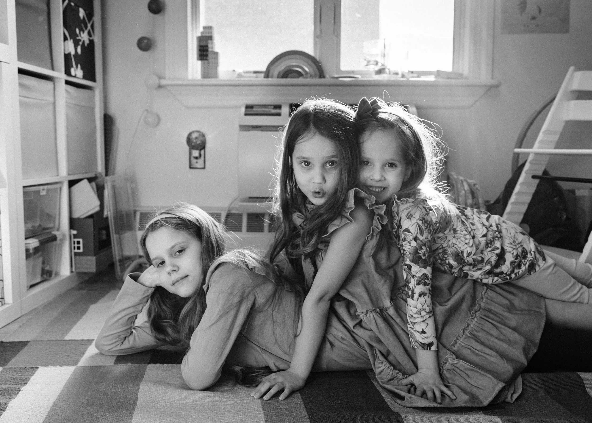 Three young girls lying on a bed in a room, posing playfully for the camera, with a window and indoor decor visible in the background.
