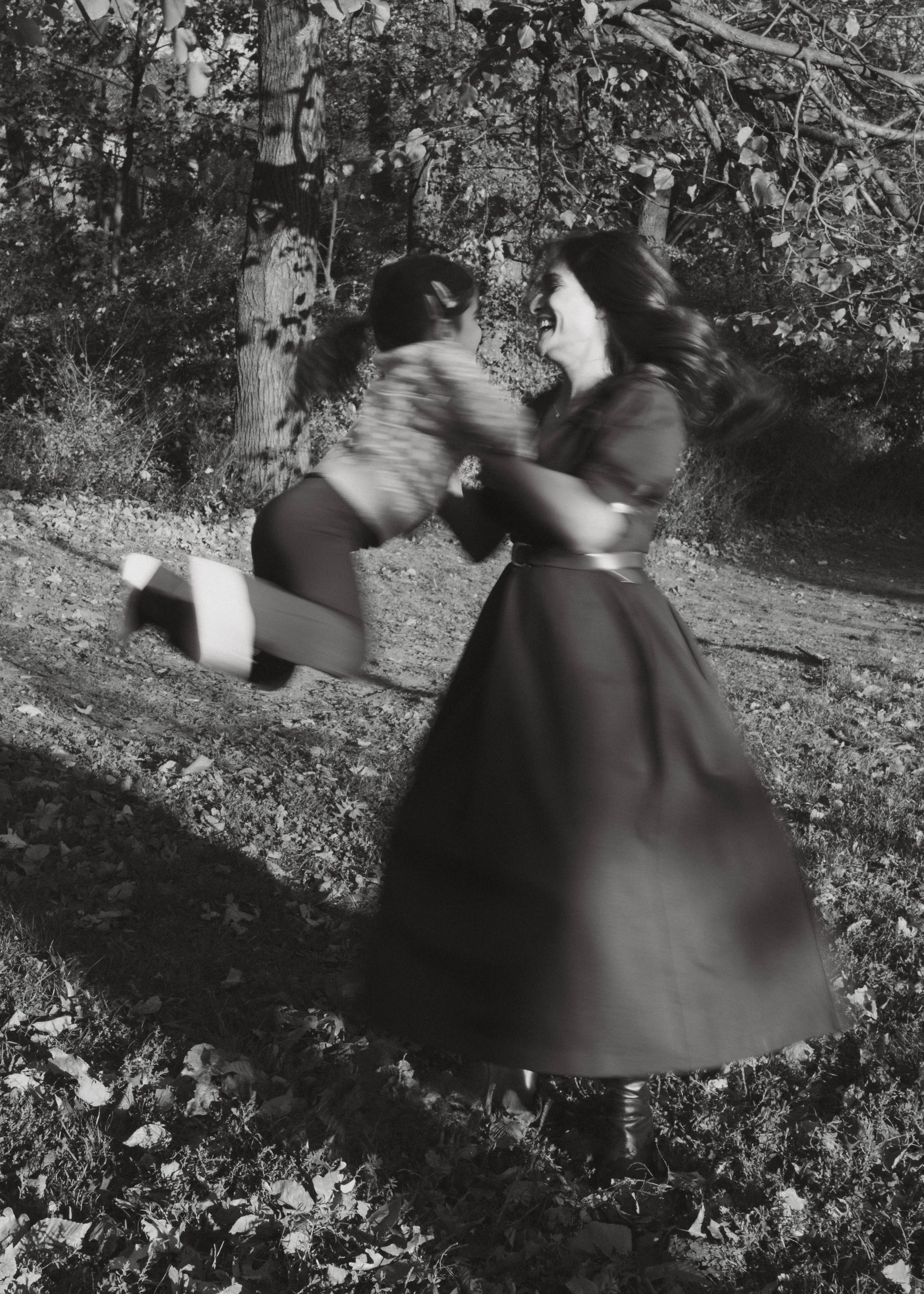 A woman in a long dark dress spinning around with a young girl in an outdoor setting with trees, laughing together at night.