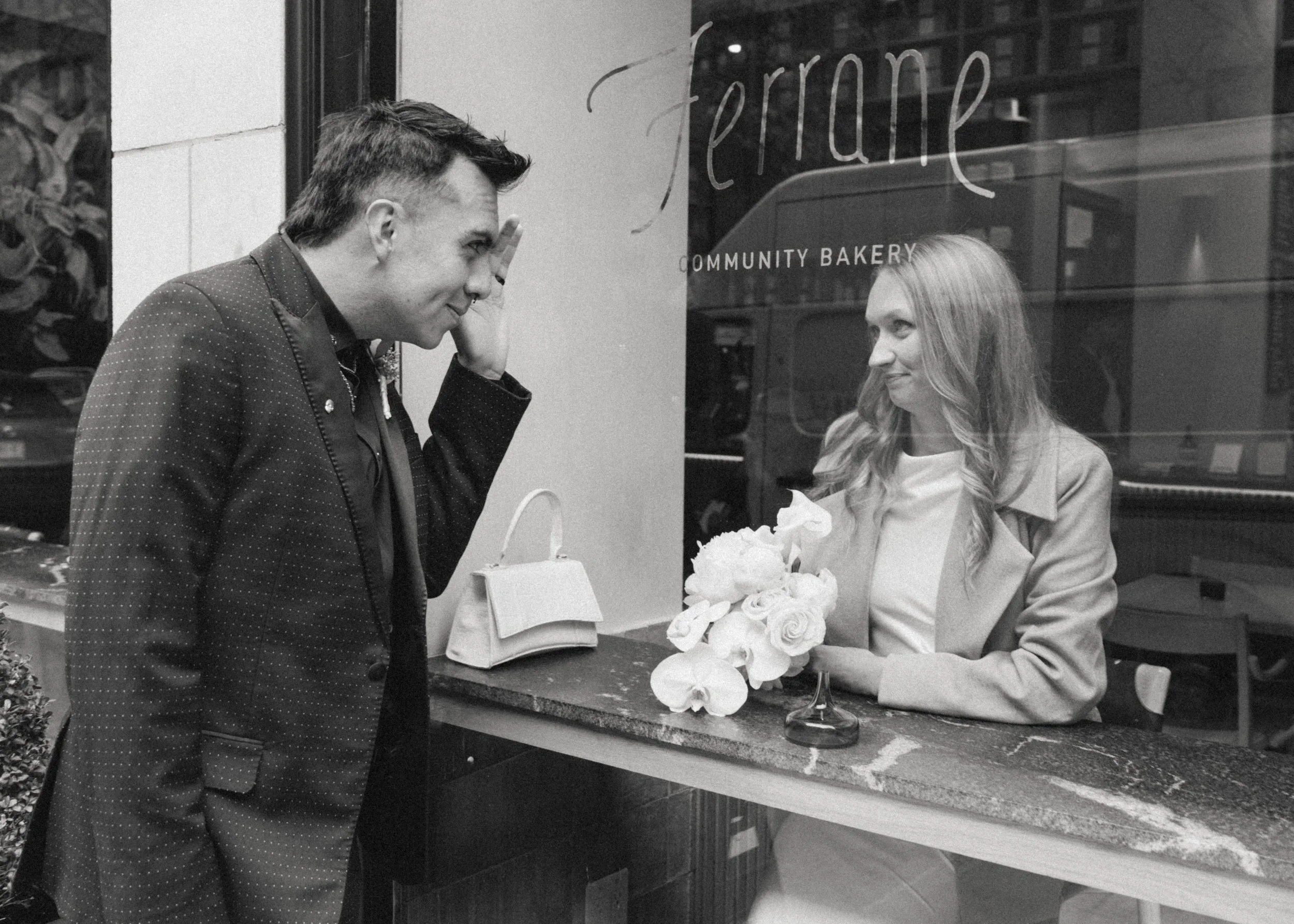 A man and a woman are interacting through a bakery window. The man is standing outside, smiling and gesturing with his hand, while the woman is inside a bakery, holding a bouquet of flowers and smiling back at him. The bakery window has the text "fer
