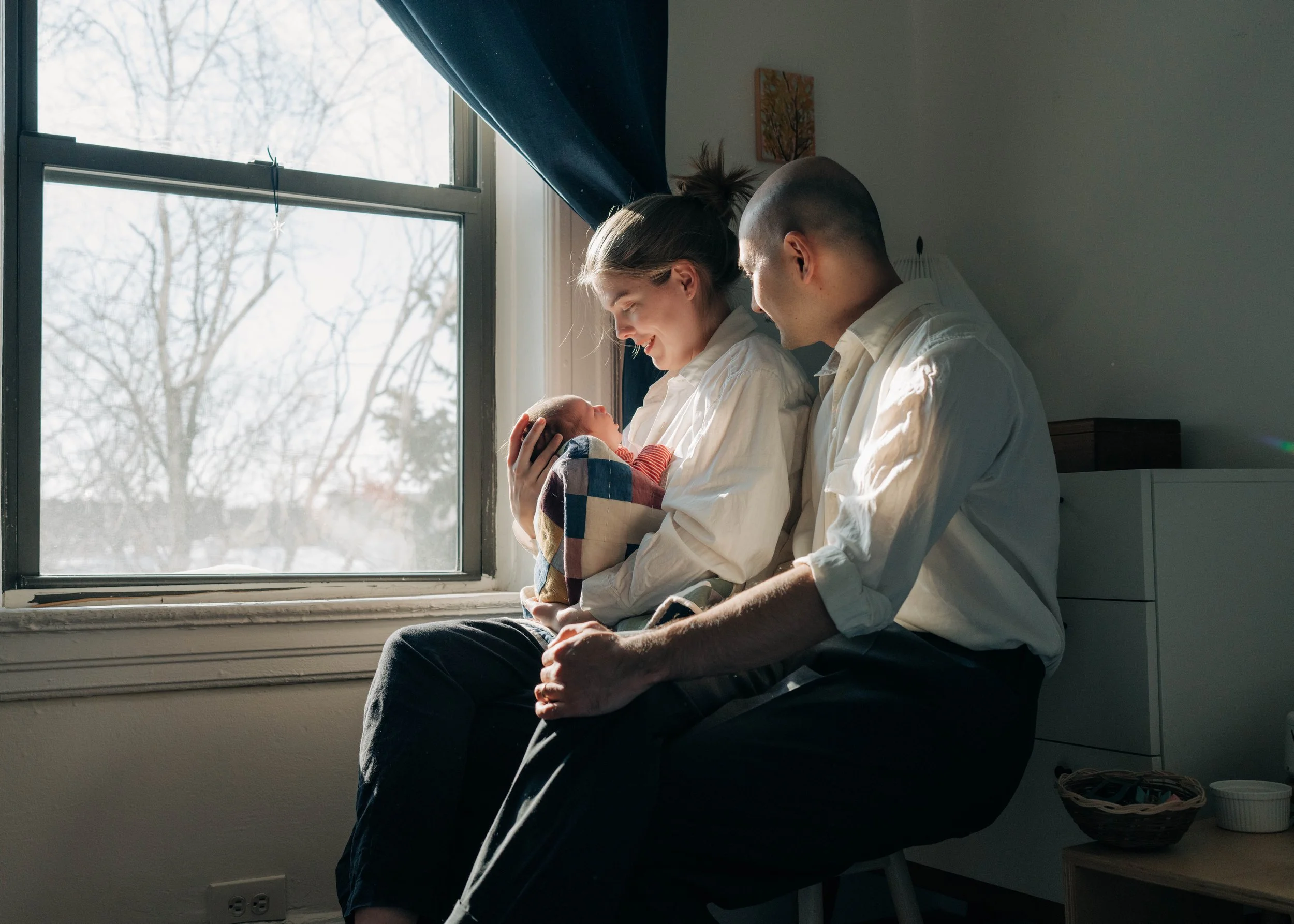 A woman holding a newborn baby is sitting on a chair with a man, by a window on a bright day.