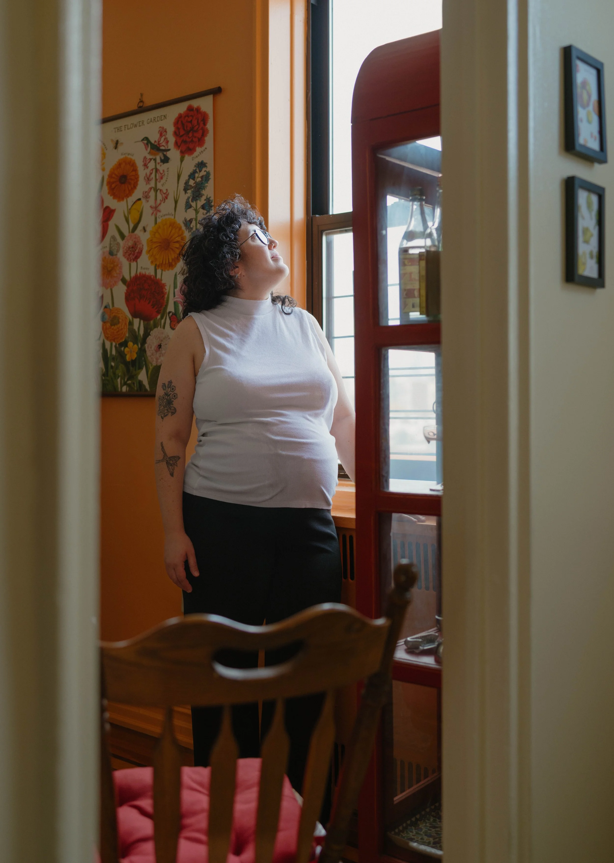 Woman with curly hair, wearing glasses, a sleeveless white top, and black pants, standing near a window in a colorful room with floral wall art.