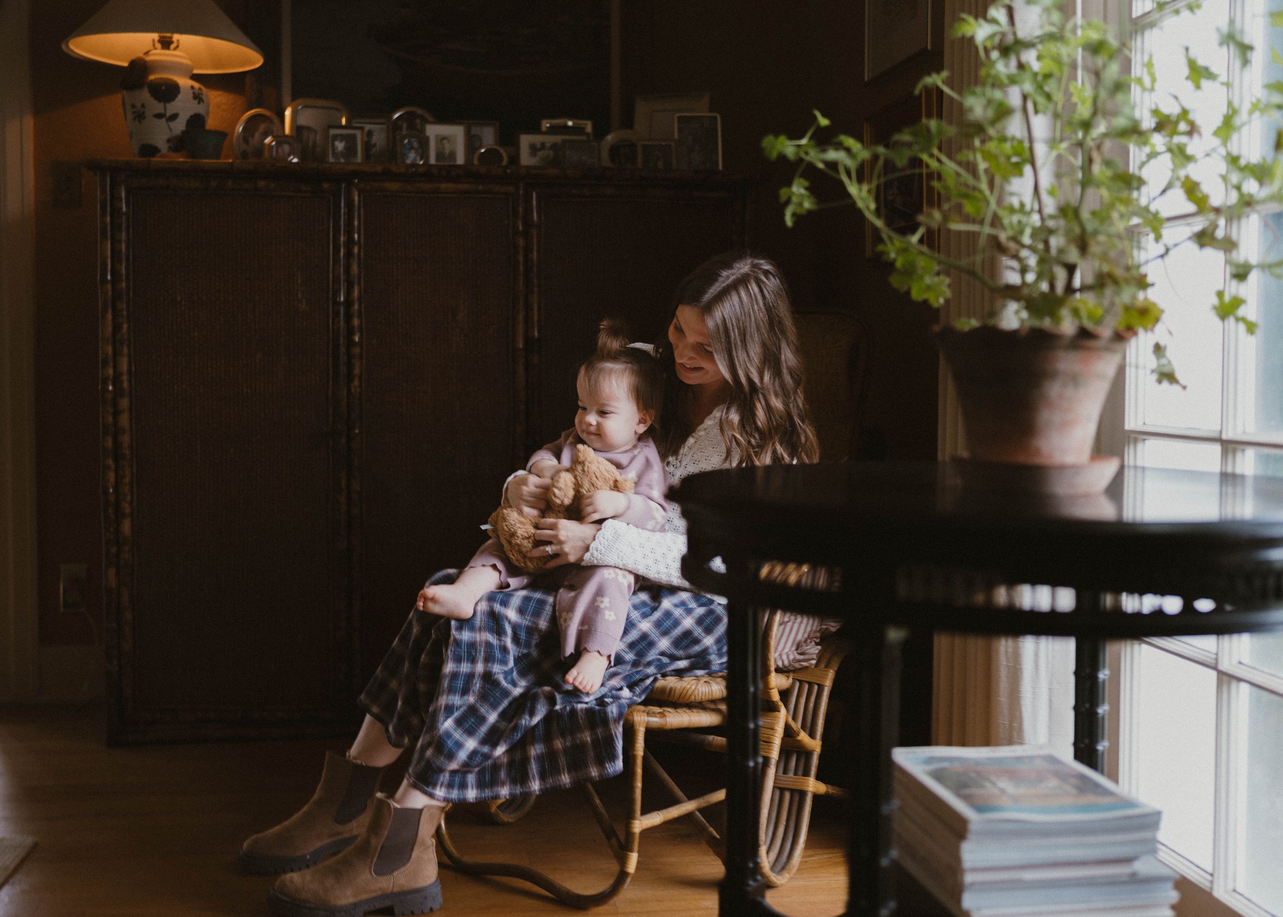 A woman with long brown hair sitting on a wooden chair, holding a young girl with front hair tied in a small ponytail. The girl is holding a teddy bear. They are in a cozy room with a large window and a potted plant. There is a stack of magazines or books in the foreground.