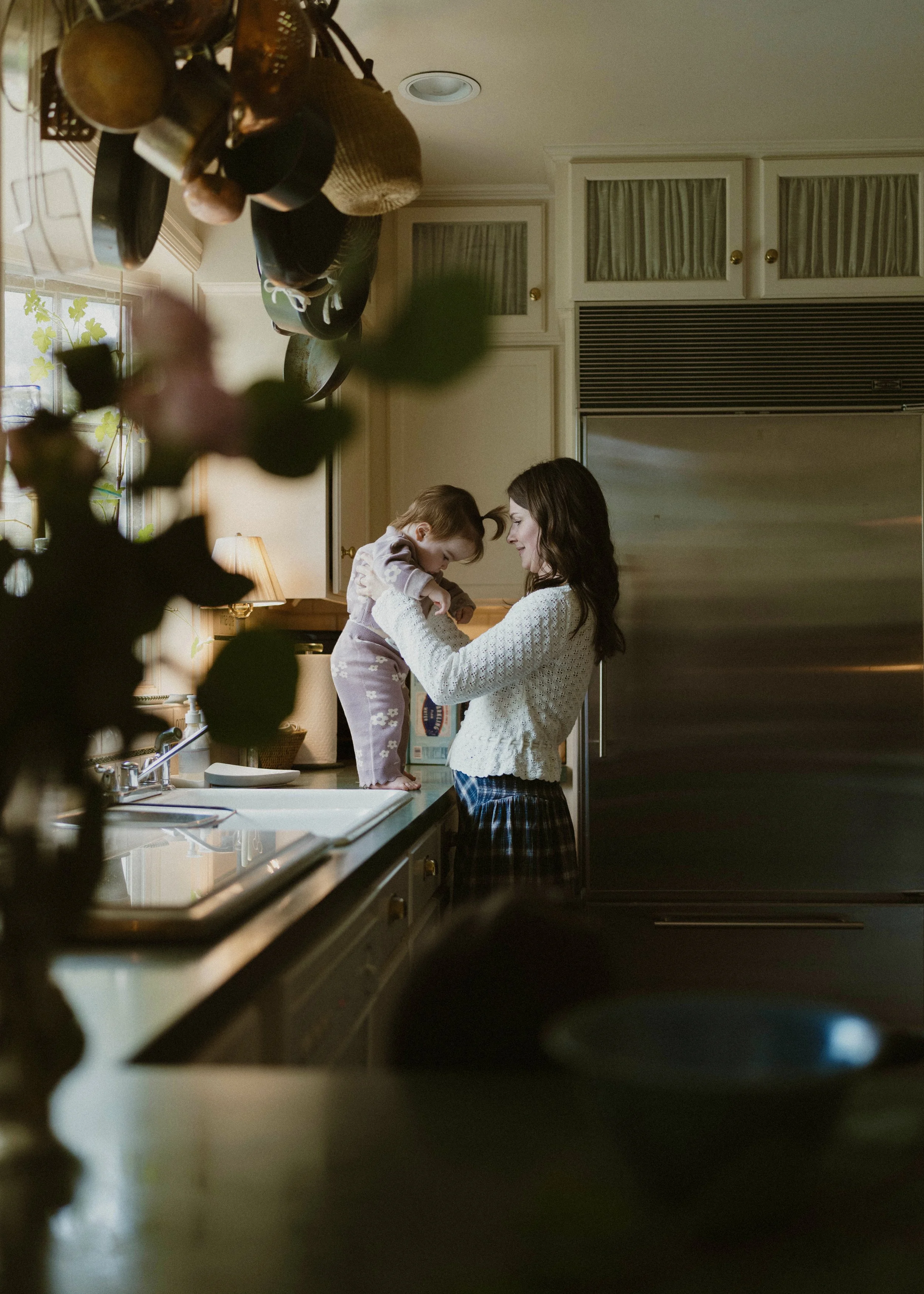 A woman with dark hair and a child standing on a kitchen counter, with the woman holding the child and both smiling. The kitchen has cabinets, a window with a plant, a lamp, and various kitchen utensils hanging from above.
