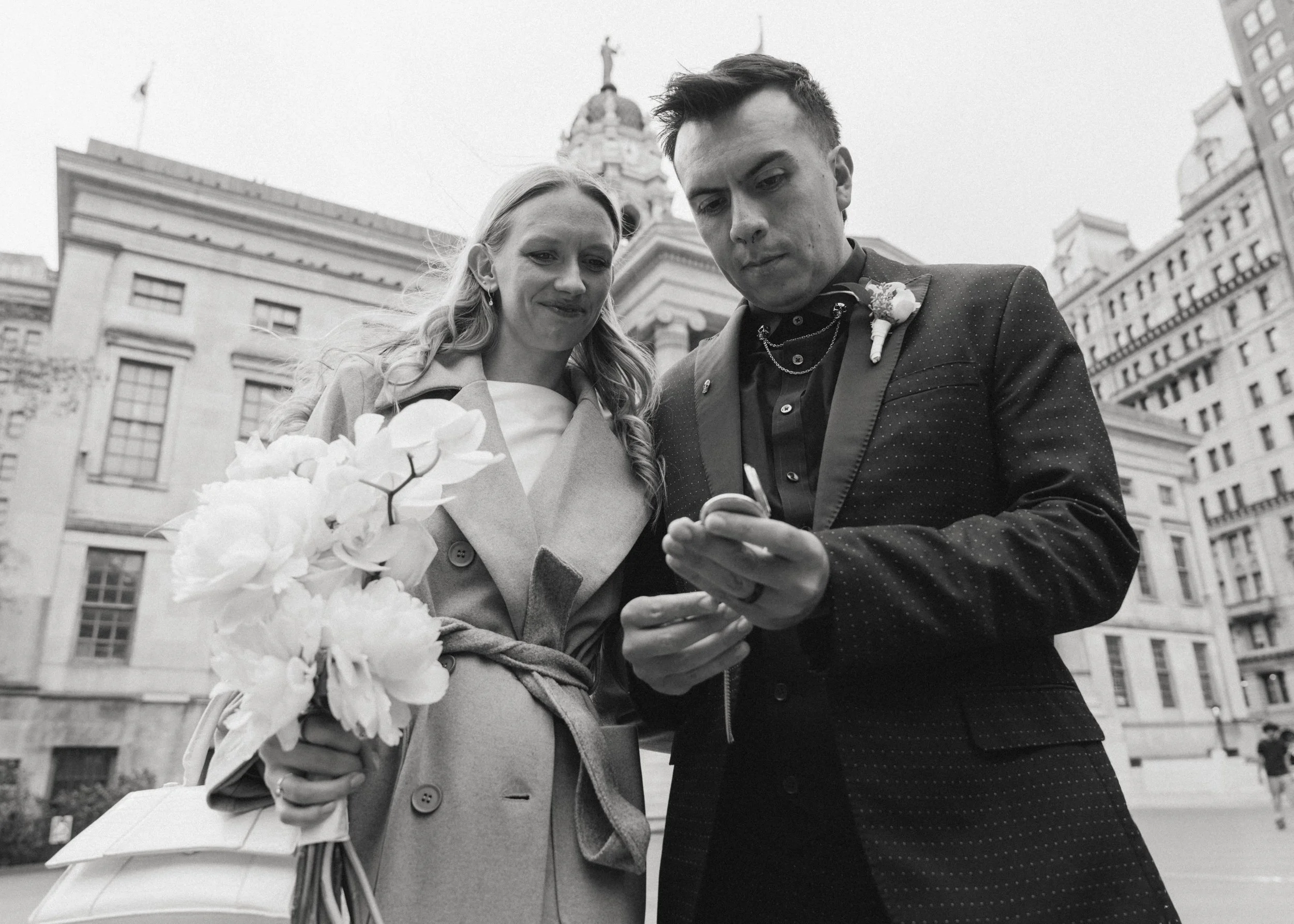 A black and white photo of a man and woman standing outdoors in a city, both looking at a smartphone. The woman holds a bouquet of white flowers and wears a light coat. The man wears a dark jacket with a boutonniere.