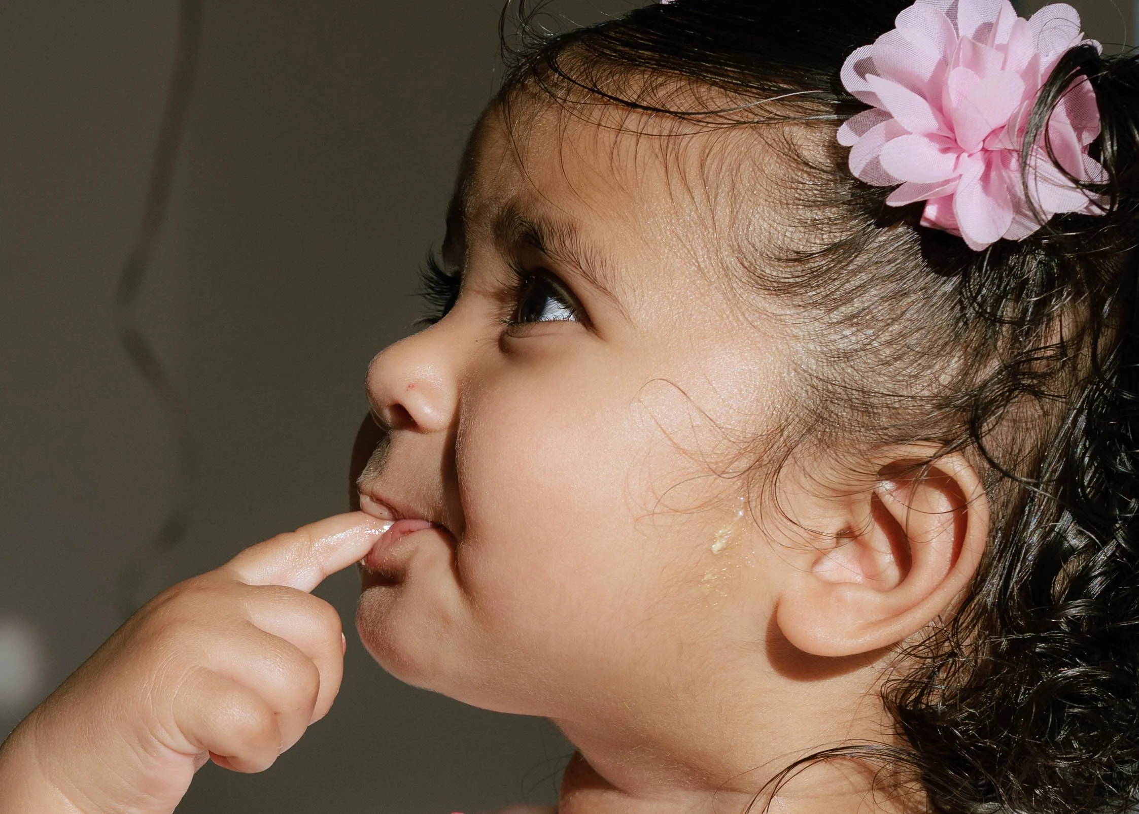 Close-up side profile of a young girl with curly dark hair, wearing a pink flower hair accessory, touching her lips with her finger, and looking upward.