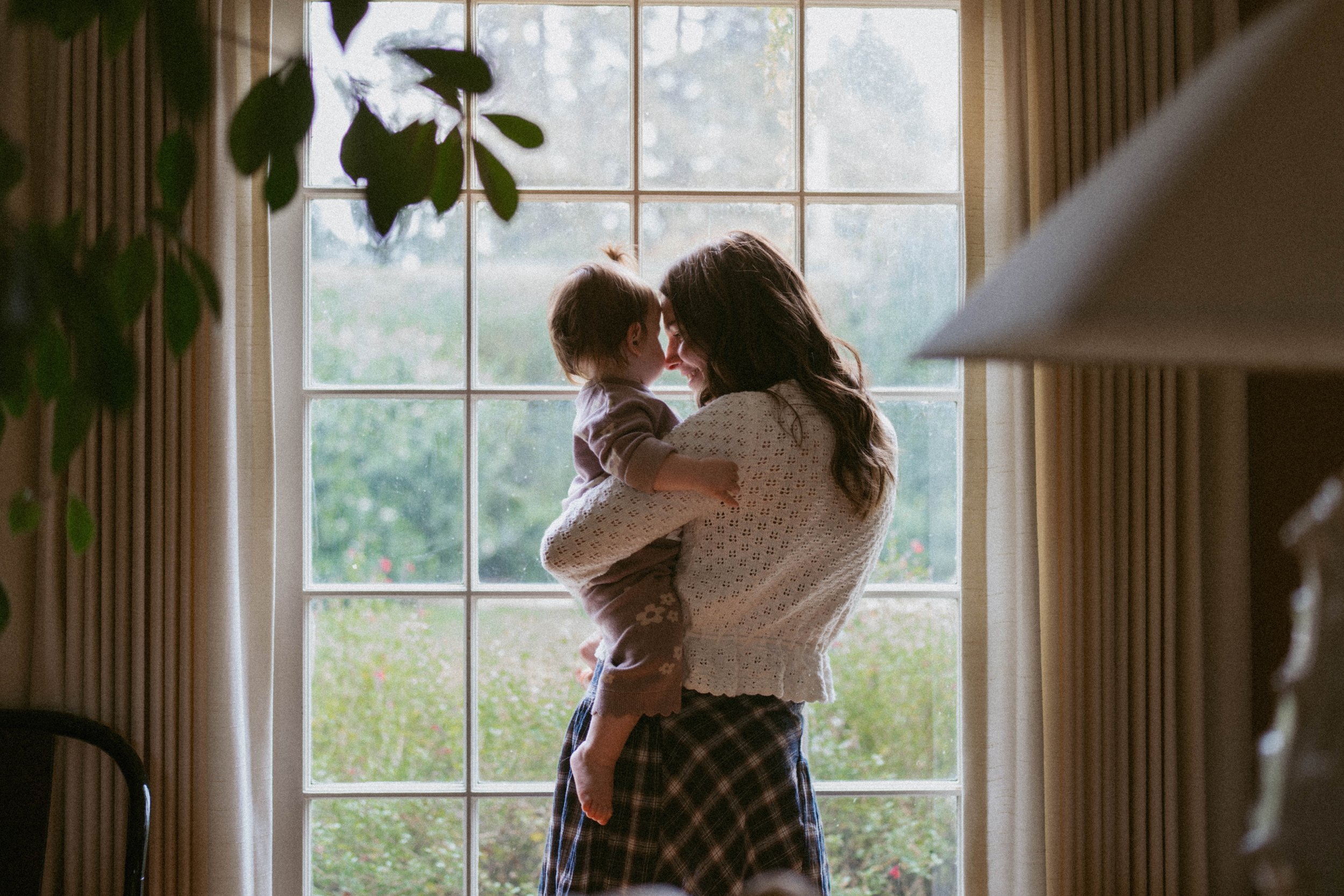 Sonja Malic captures a cinematic image of a woman holding a young child in front of a window with curtains, outdoors visible through the window.