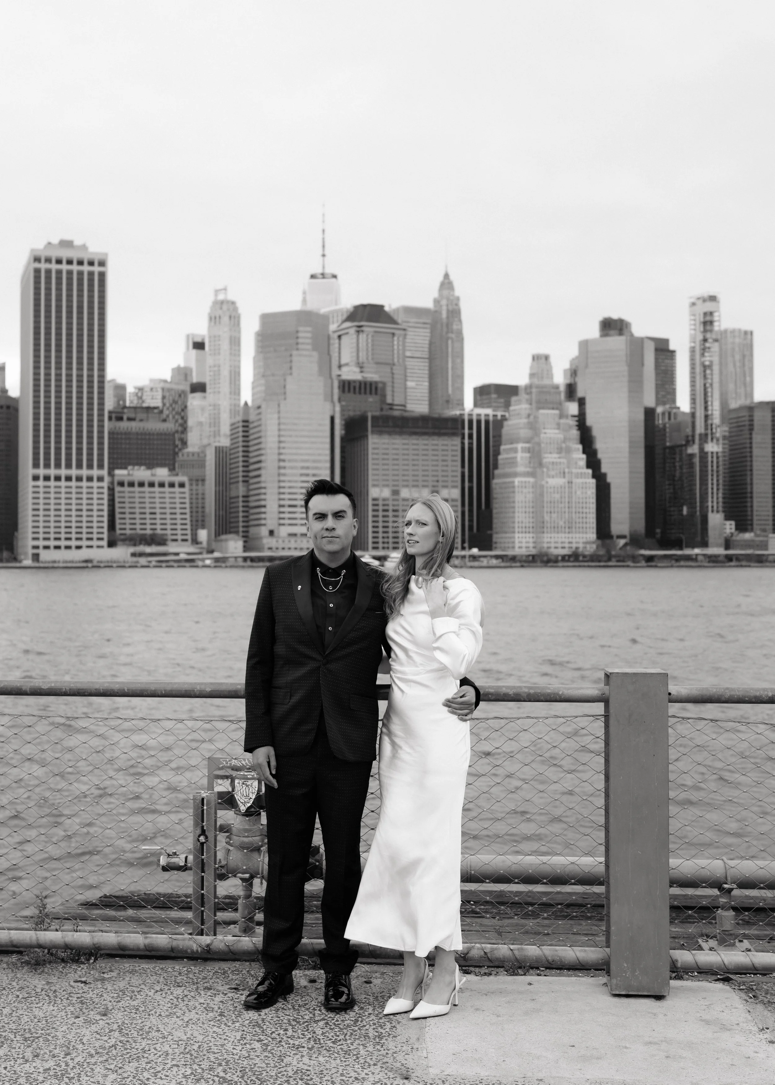 A black-and-white photo of a man and a woman standing by a waterfront with New York City’s skyline in the background. The man is dressed in a dark suit with jewelry, and the woman is wearing a long white dress with high heels.