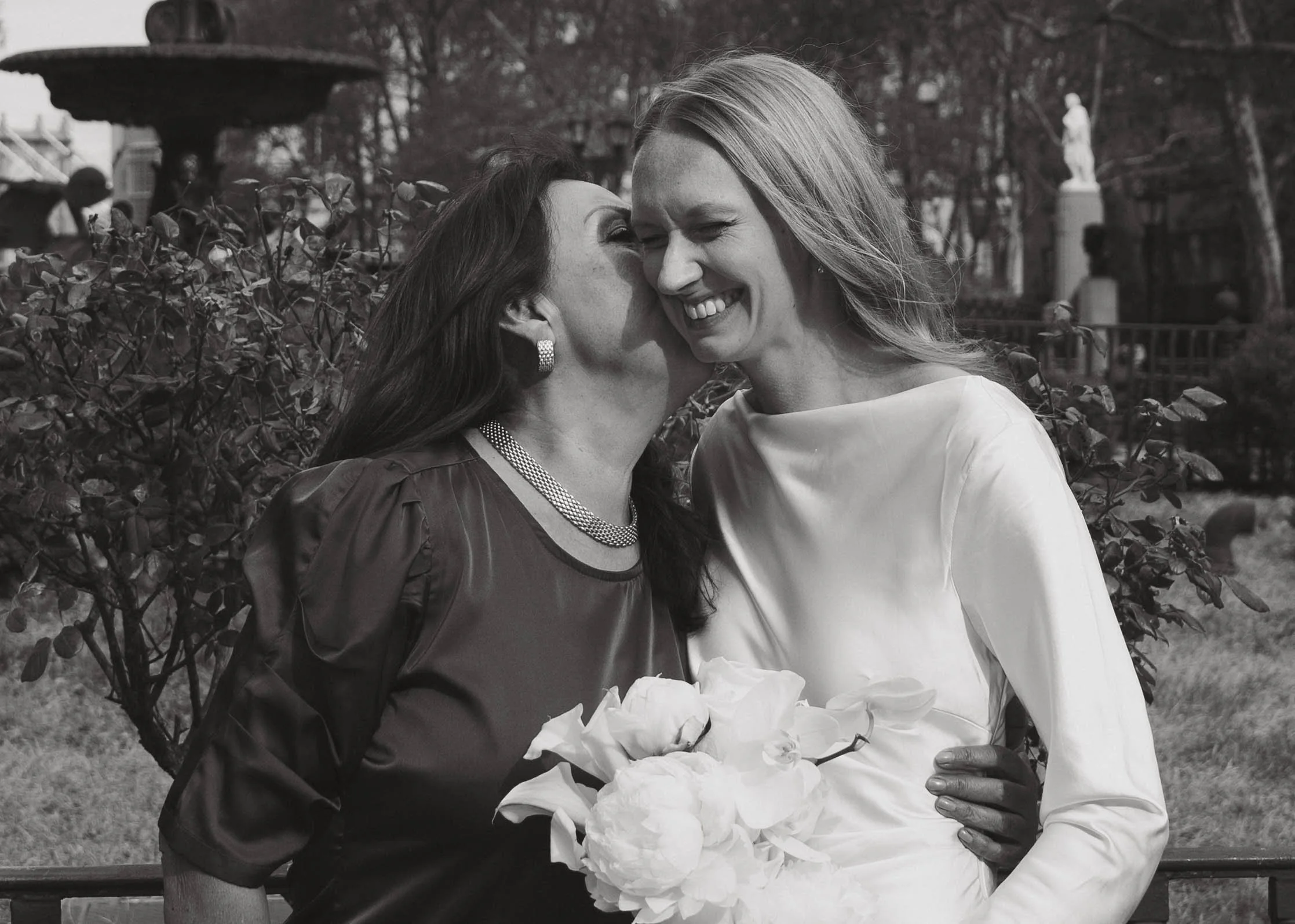 Two women share a joyful moment outdoors, one whispering into the other's ear and both smiling. The woman on the right is holding a bouquet of flowers.