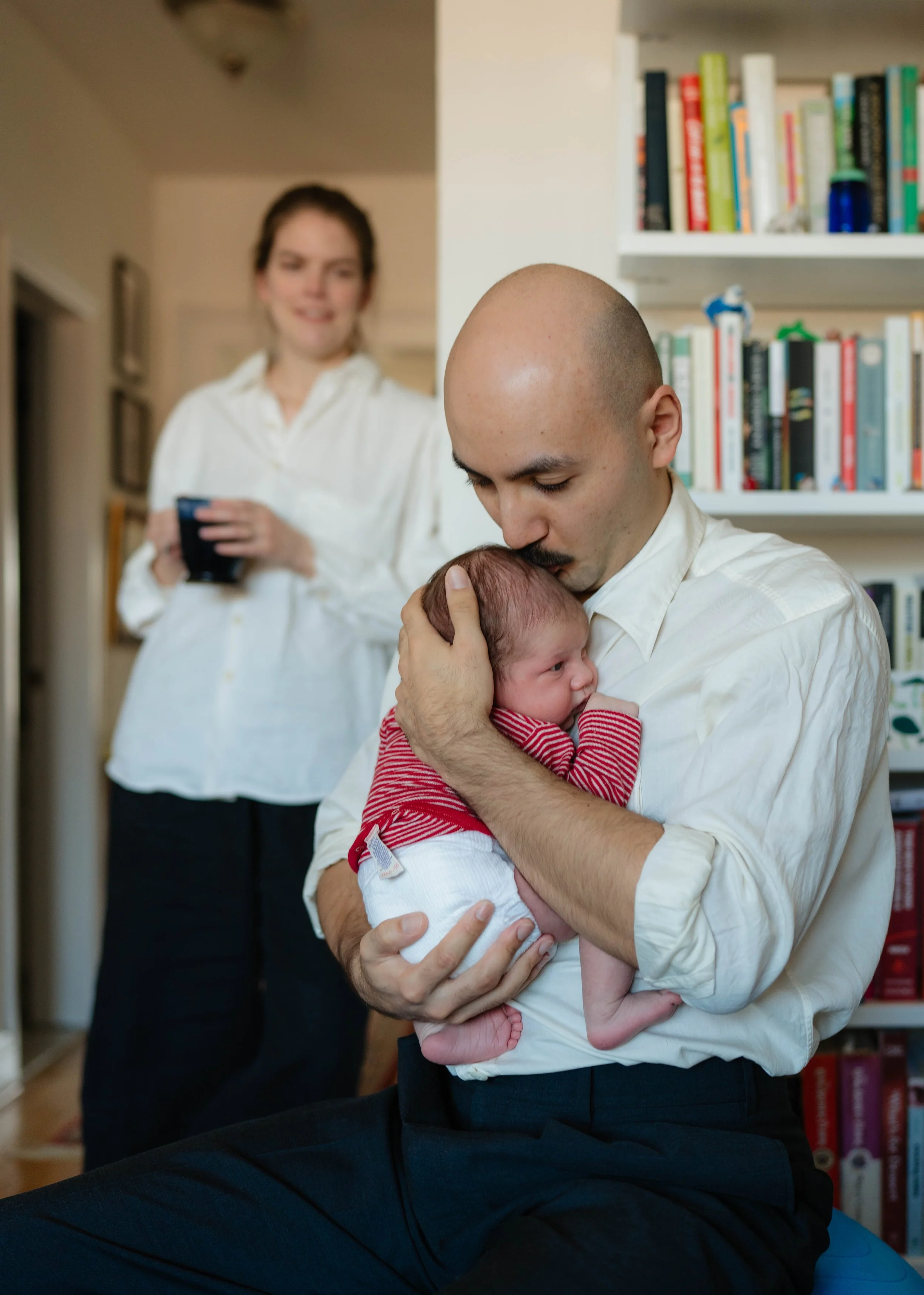 A man with a shaved head and a woman with brown hair in a white shirt are in a home, with a bookshelf in the background. The man is holding a baby with brown hair and a red and white striped shirt, kissing the baby's head. The woman is holding a mug 