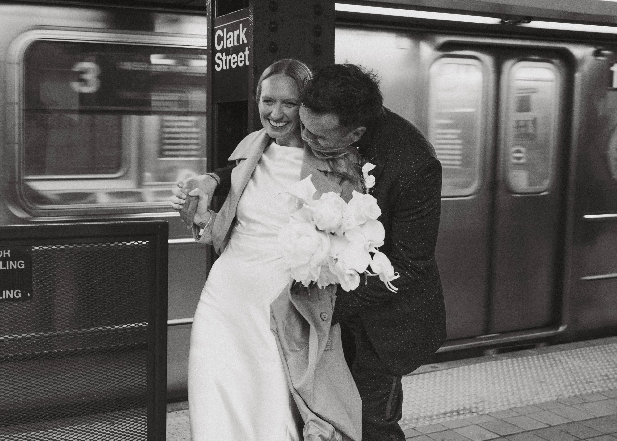 A bride and groom share a joyful moment at a subway station, with the bride holding a large bouquet of white flowers and the groom leaning in close, both smiling. The station sign reads 'Clark Street'.