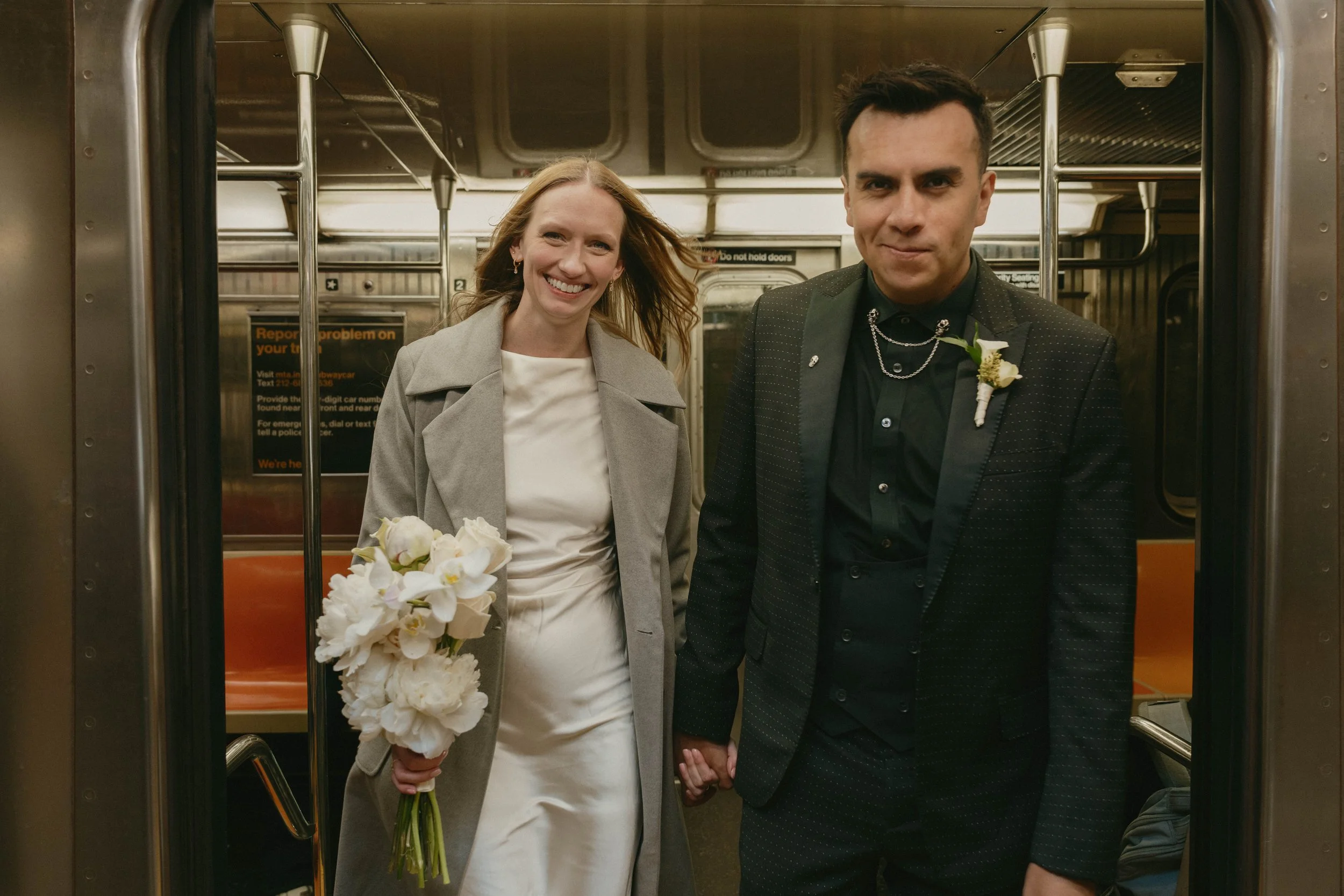 A smiling woman in a light gray coat holding a bouquet of white flowers, standing next to a man in a black tuxedo with a boutonniere, both holding hands inside a subway train.