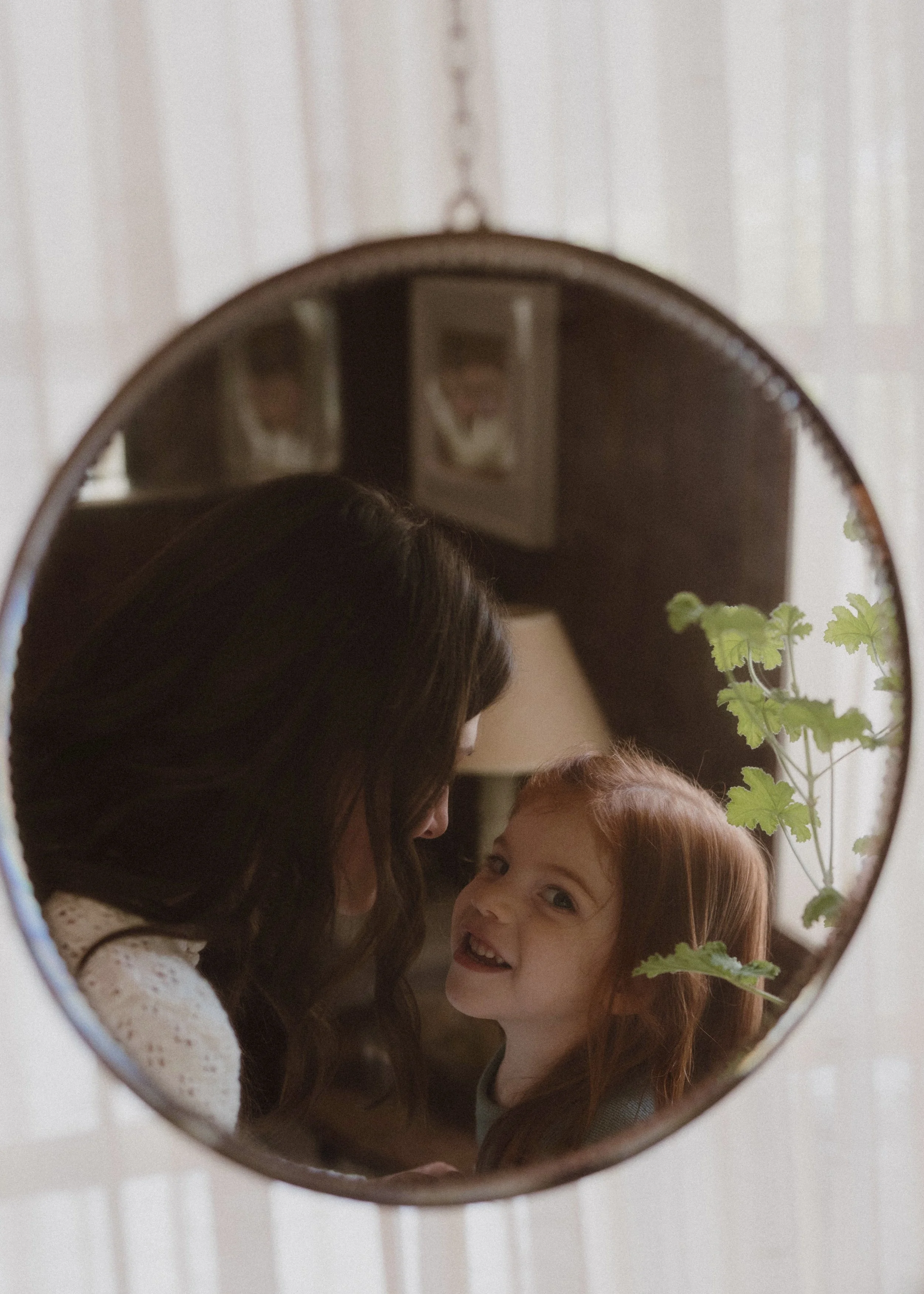 A mirror reflecting a woman and a young girl smiling and looking at each other, with a plant with green leaves in the foreground.
