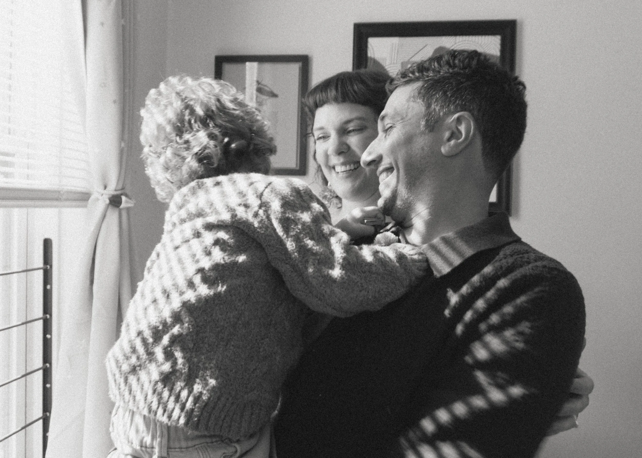 A black-and-white photo of a happy family with a young girl and their dog, sharing a joyful moment inside a home with framed pictures on the wall.