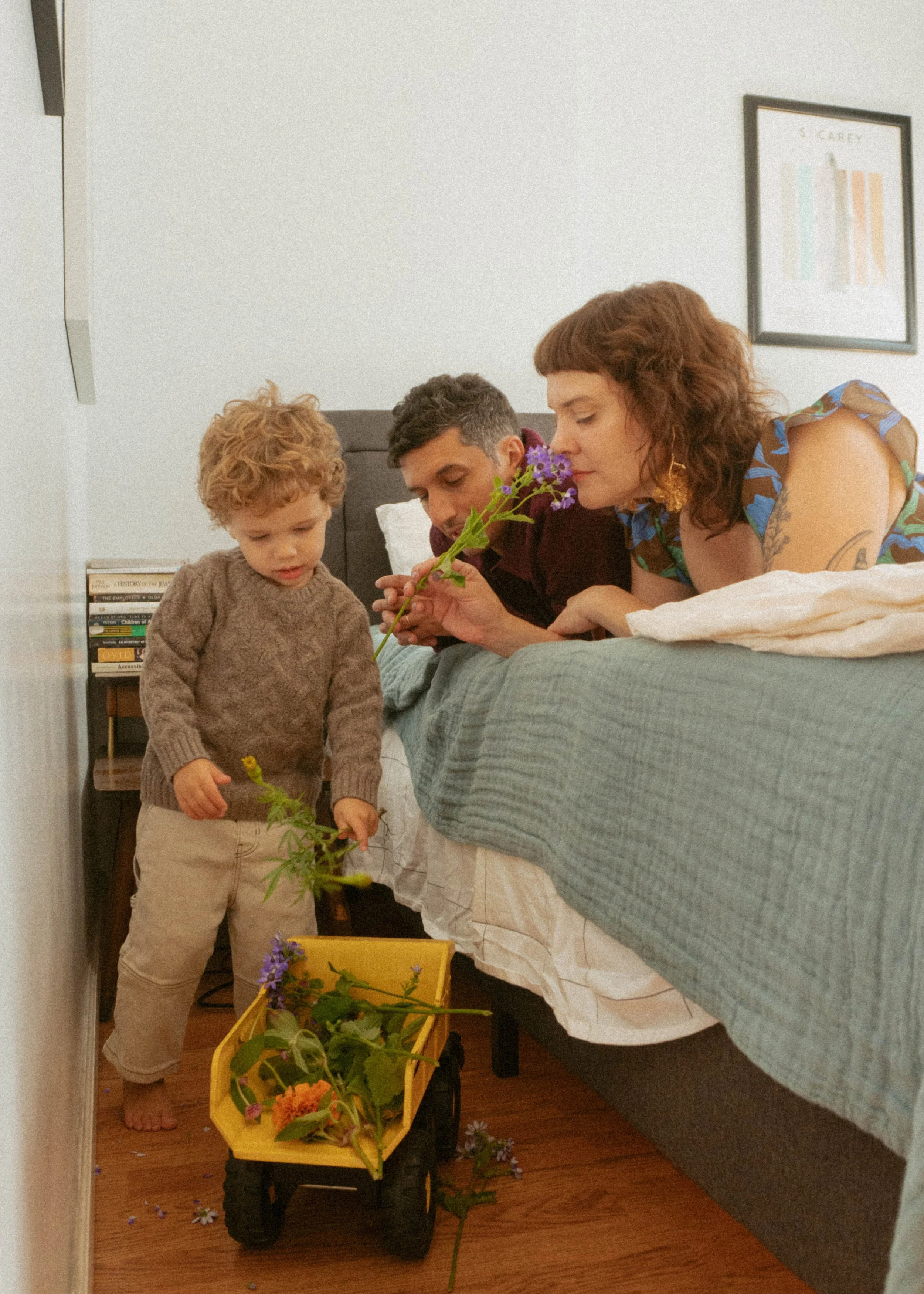 Family on a bed playing with colorful flowers in a yellow wagon, with a bookshelf and framed art on the wall in the background.