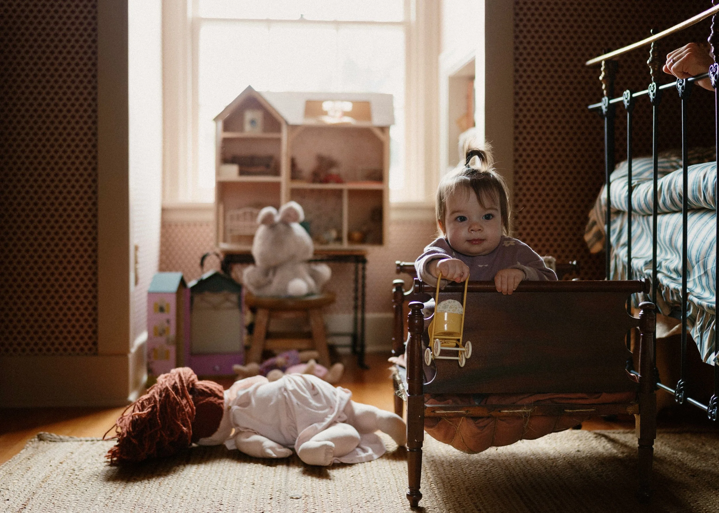 A young girl with a ponytail is sitting inside a bed's frame, holding a yellow toy shopping cart, in a sunlit bedroom with scattered stuffed animals and toys on the floor.