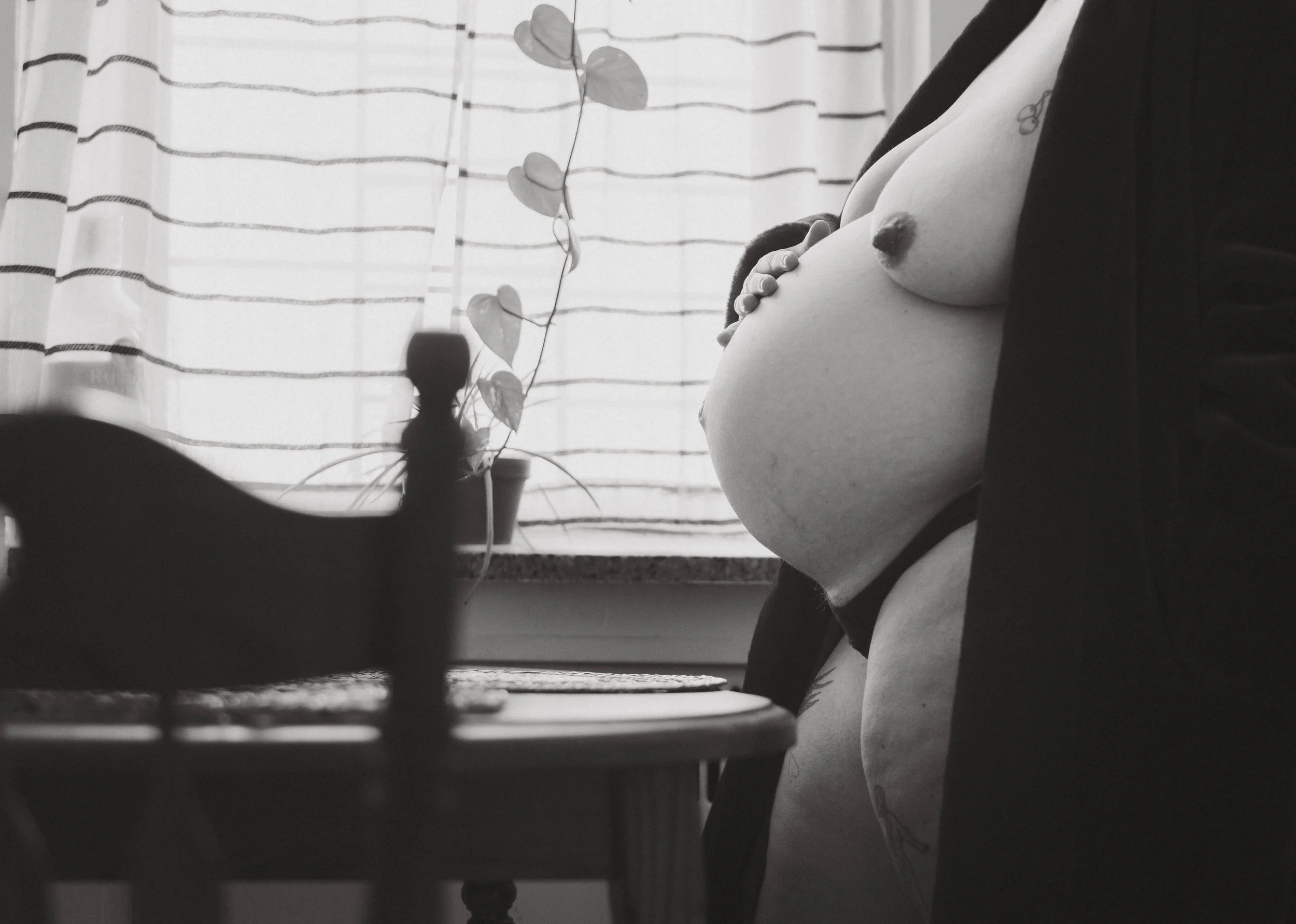 A black and white photo of a pregnant woman standing next to a table with a small plant and curtain in the background.