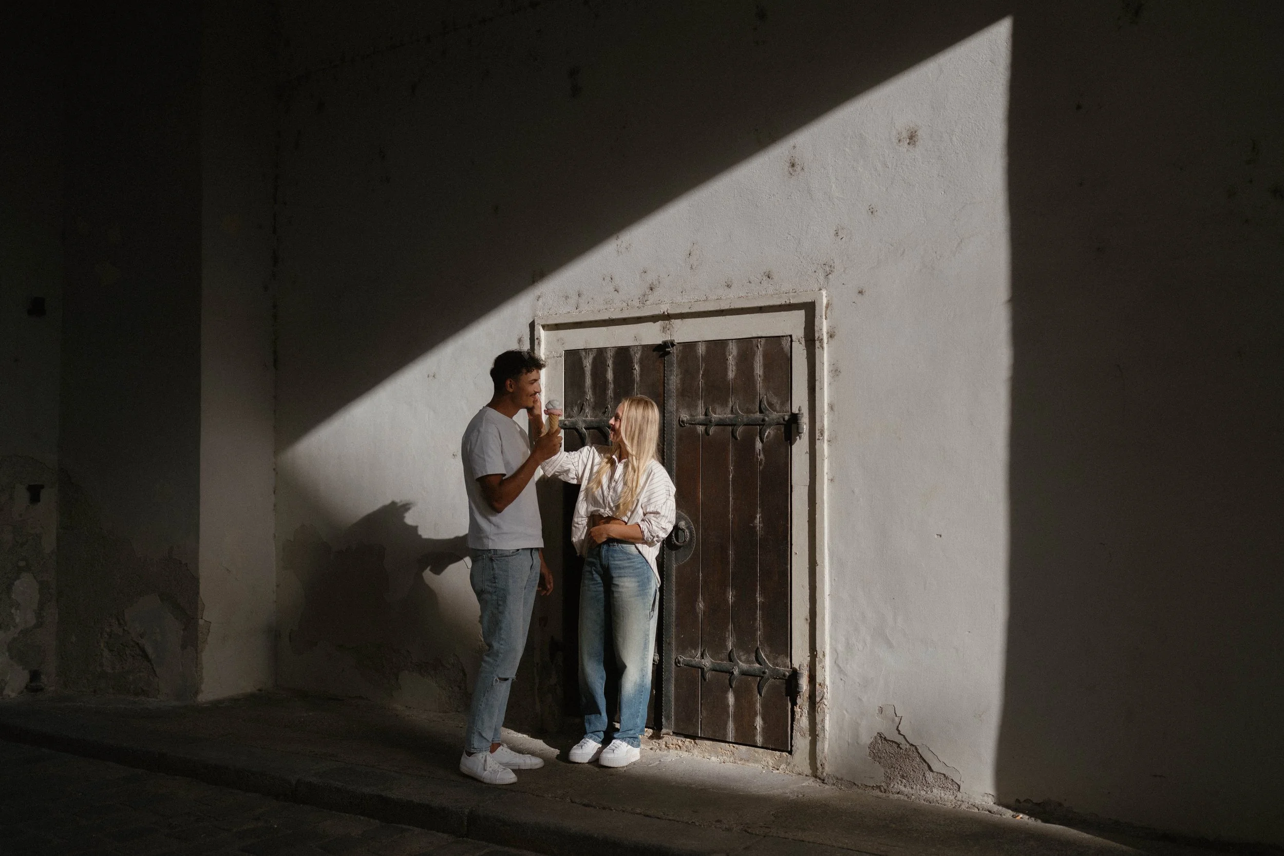 A young man and woman stand close together outside a white wall with a wooden door, sharing a moment while holding ice cream cones, illuminated by sunlight creating stark shadows.