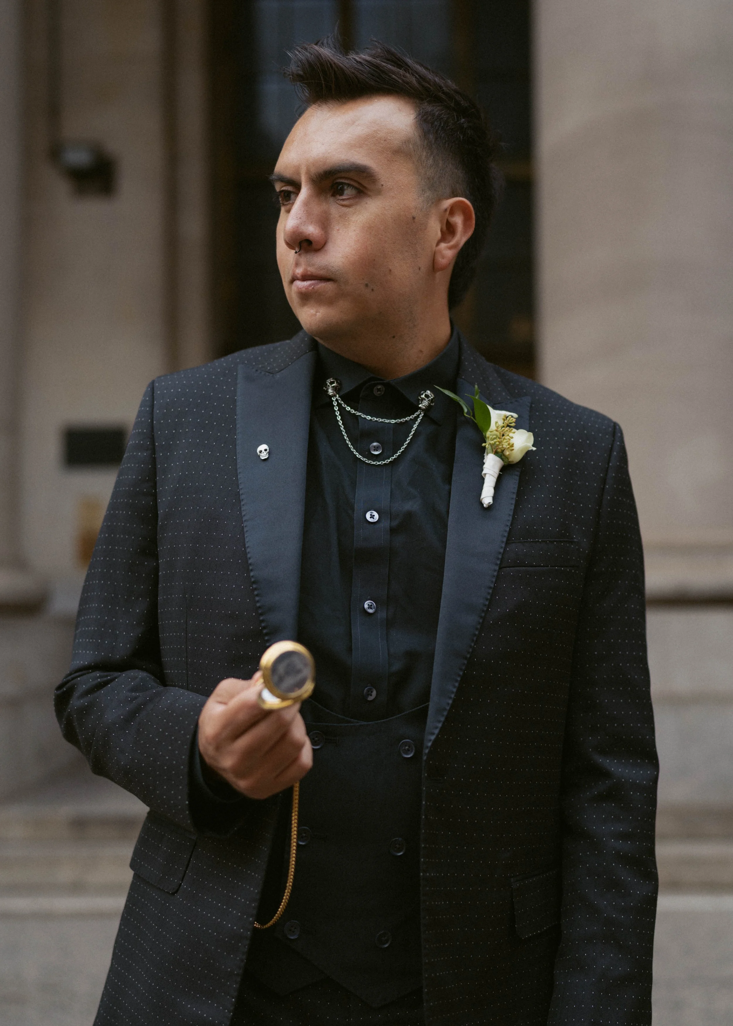 A man dressed in a black suit with subtle polka dots, black shirt, and accessories including chains, an eye pin, a skull lapel pin, and a boutonniere. He has dark hair styled upward and holds a gold pocket watch. The background appears to be an elega