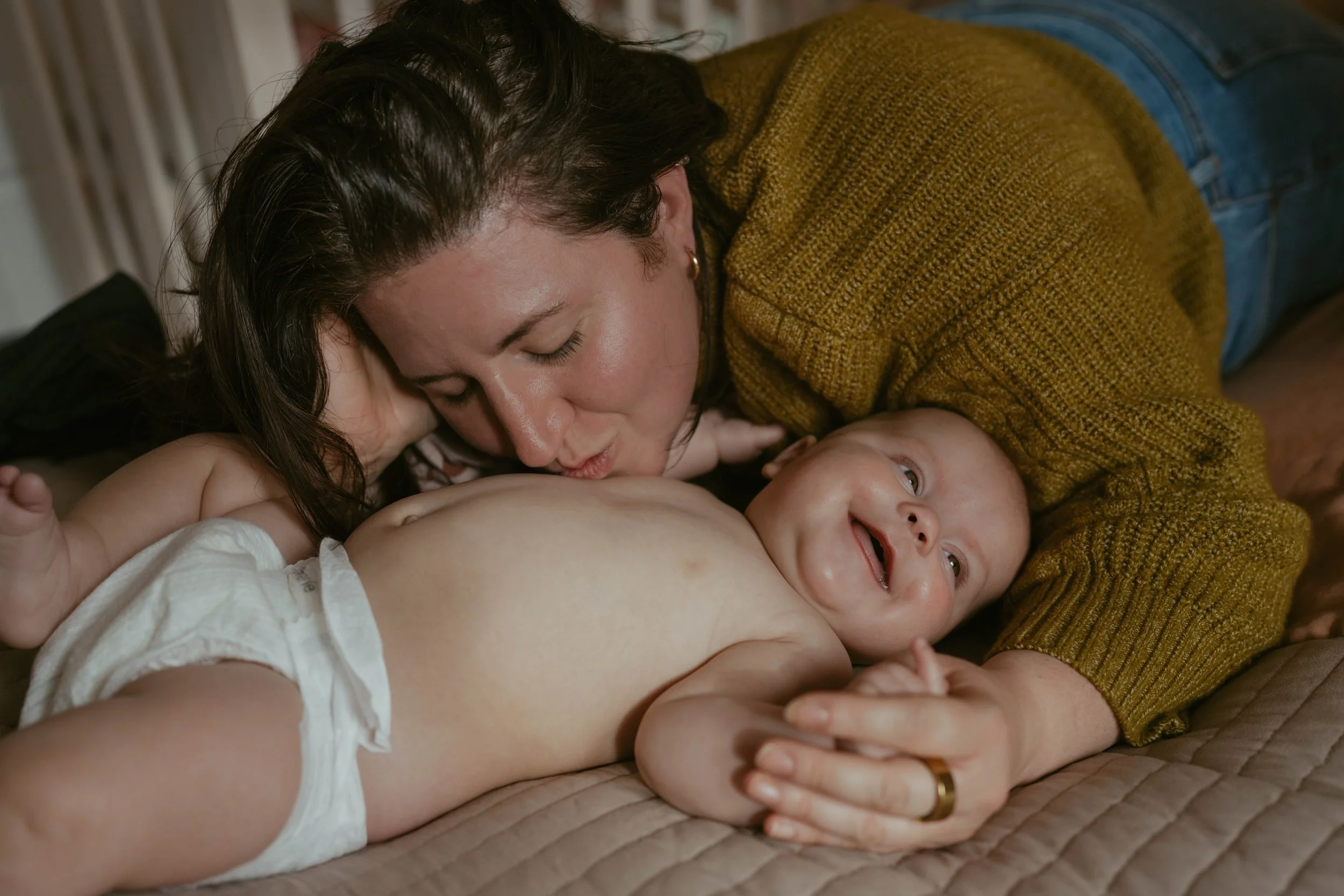 A woman is lying on a bed kissing a laughing baby who is lying on his back, wearing only a diaper. The woman has dark hair and is wearing a mustard-colored sweater. The baby has light-colored hair and eyes and is smiling.