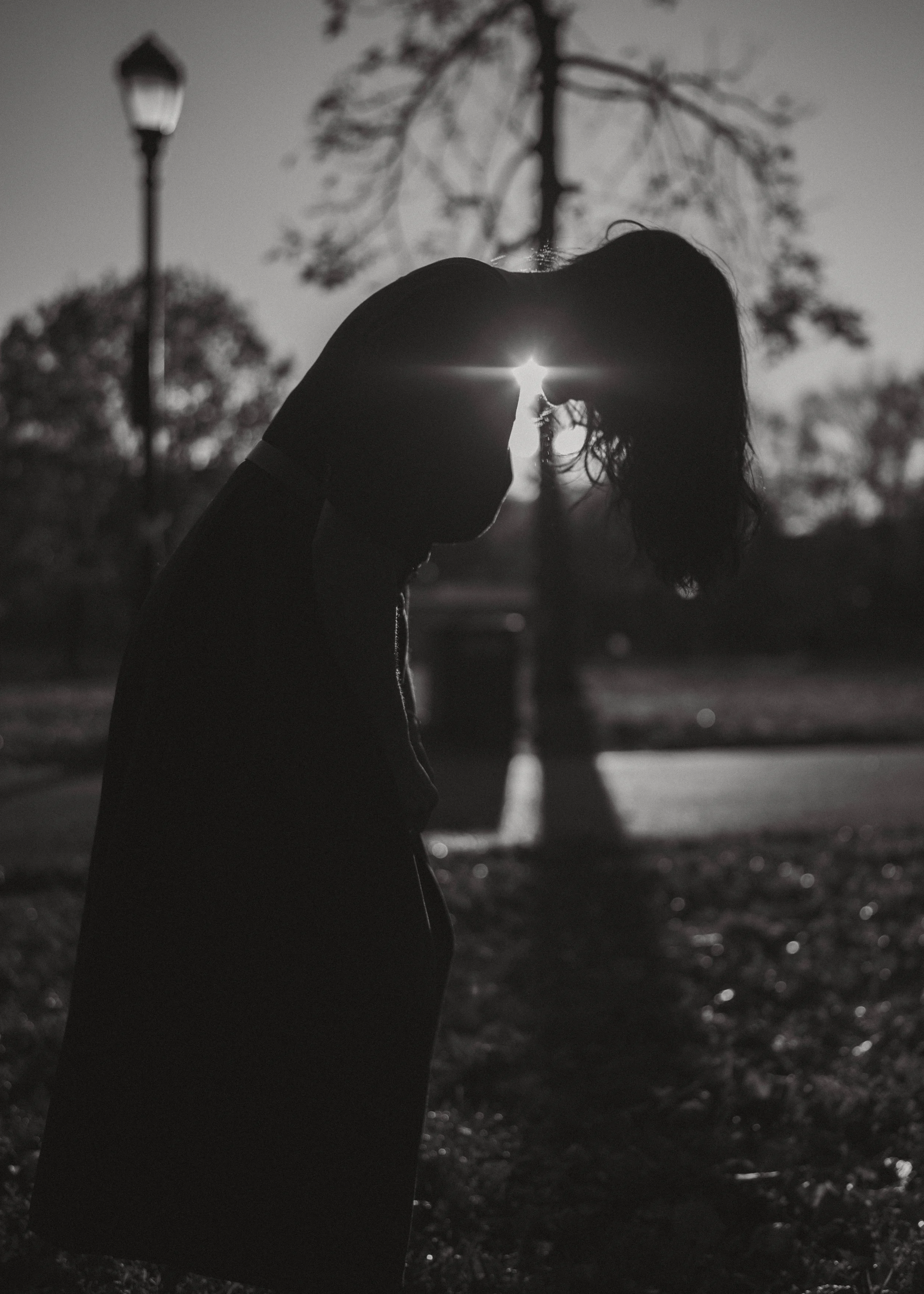 A silhouette of a woman with her head bowed, standing outdoors near a lamp post and tree, during sunset or sunrise, in black and white.