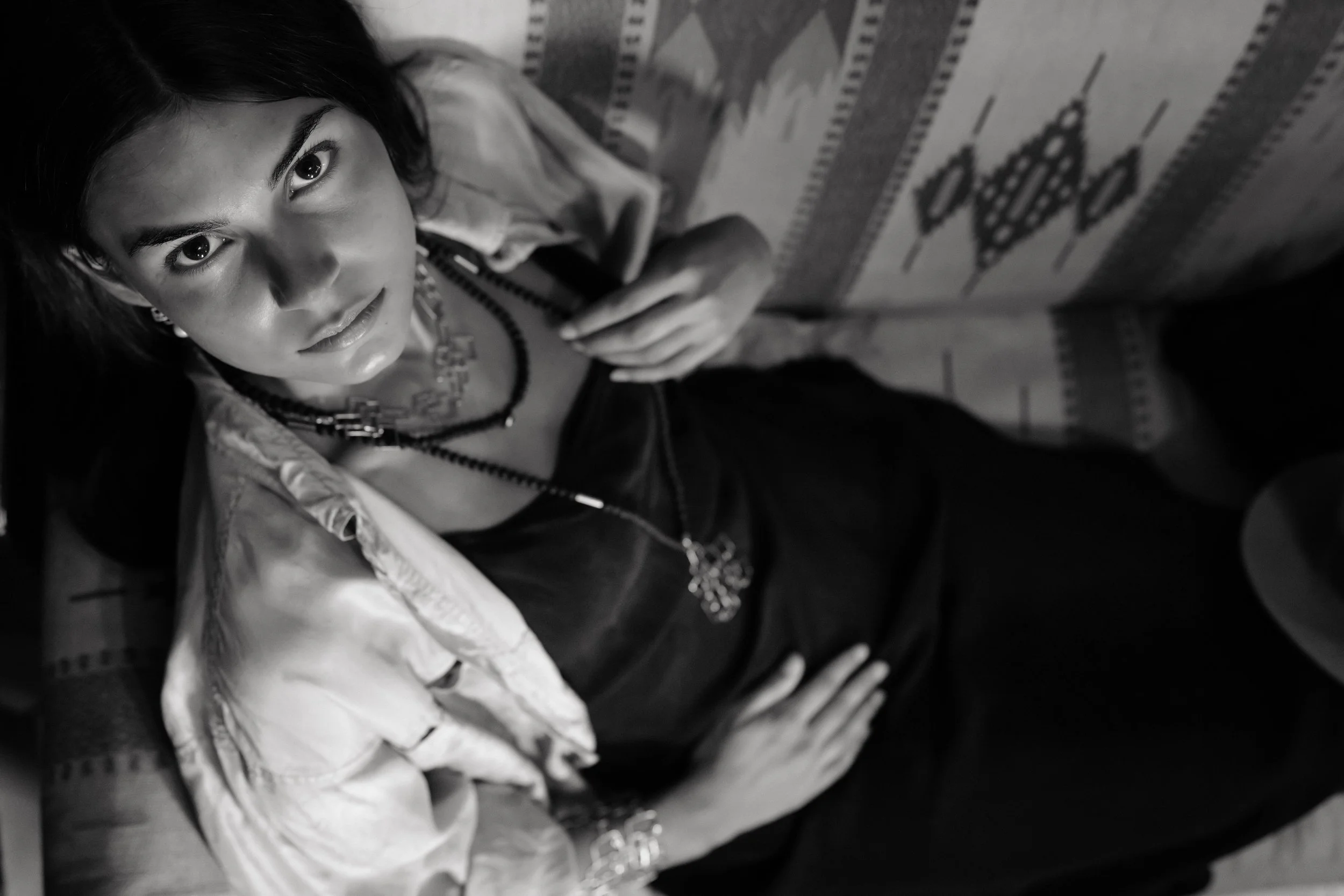 A woman with dark hair and jewelry, lying on a decorative patterned surface, looking up at the camera.