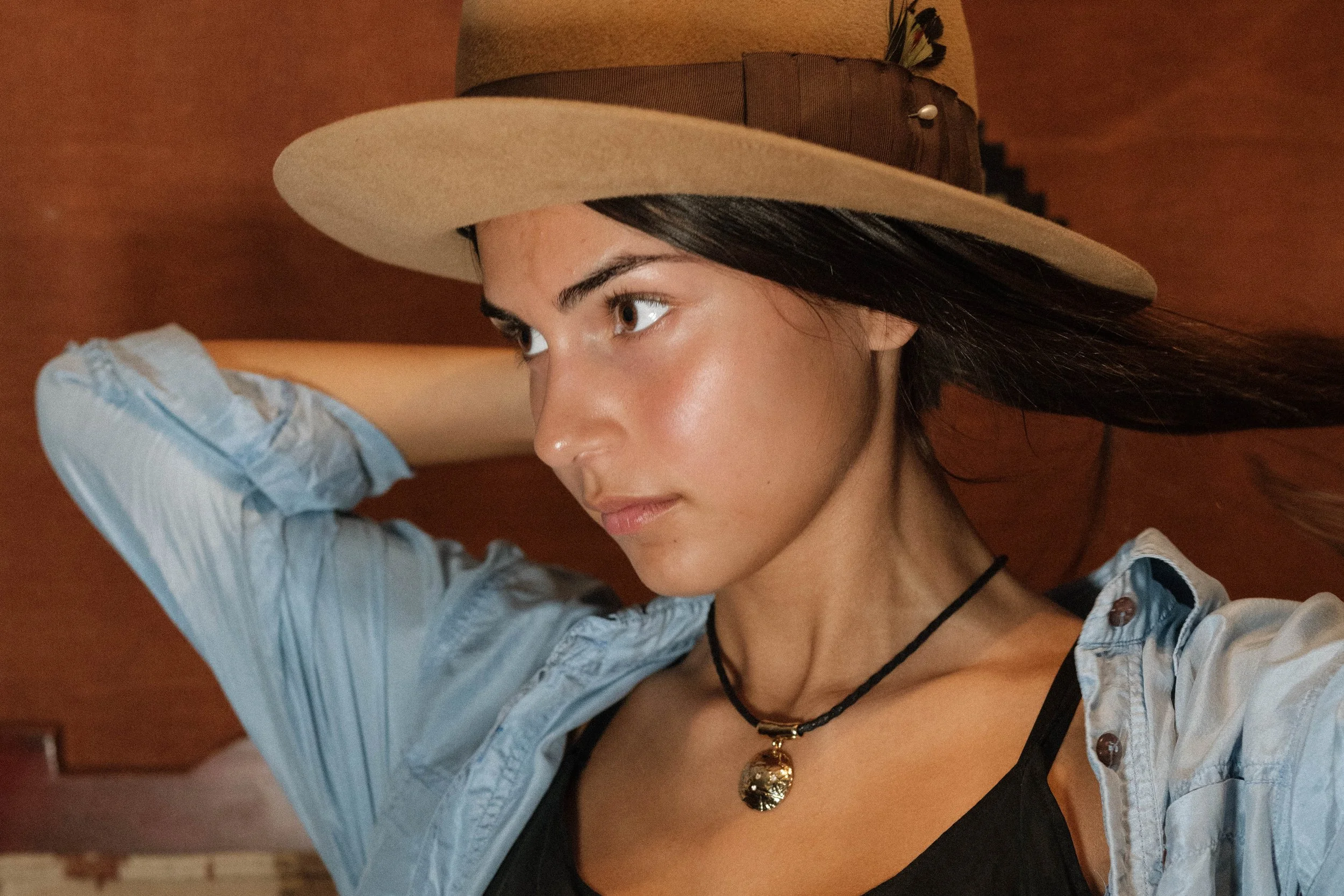 Woman wearing a wide-brimmed hat, a blue button-up shirt, a black top, and a black necklace with a gold pendant.