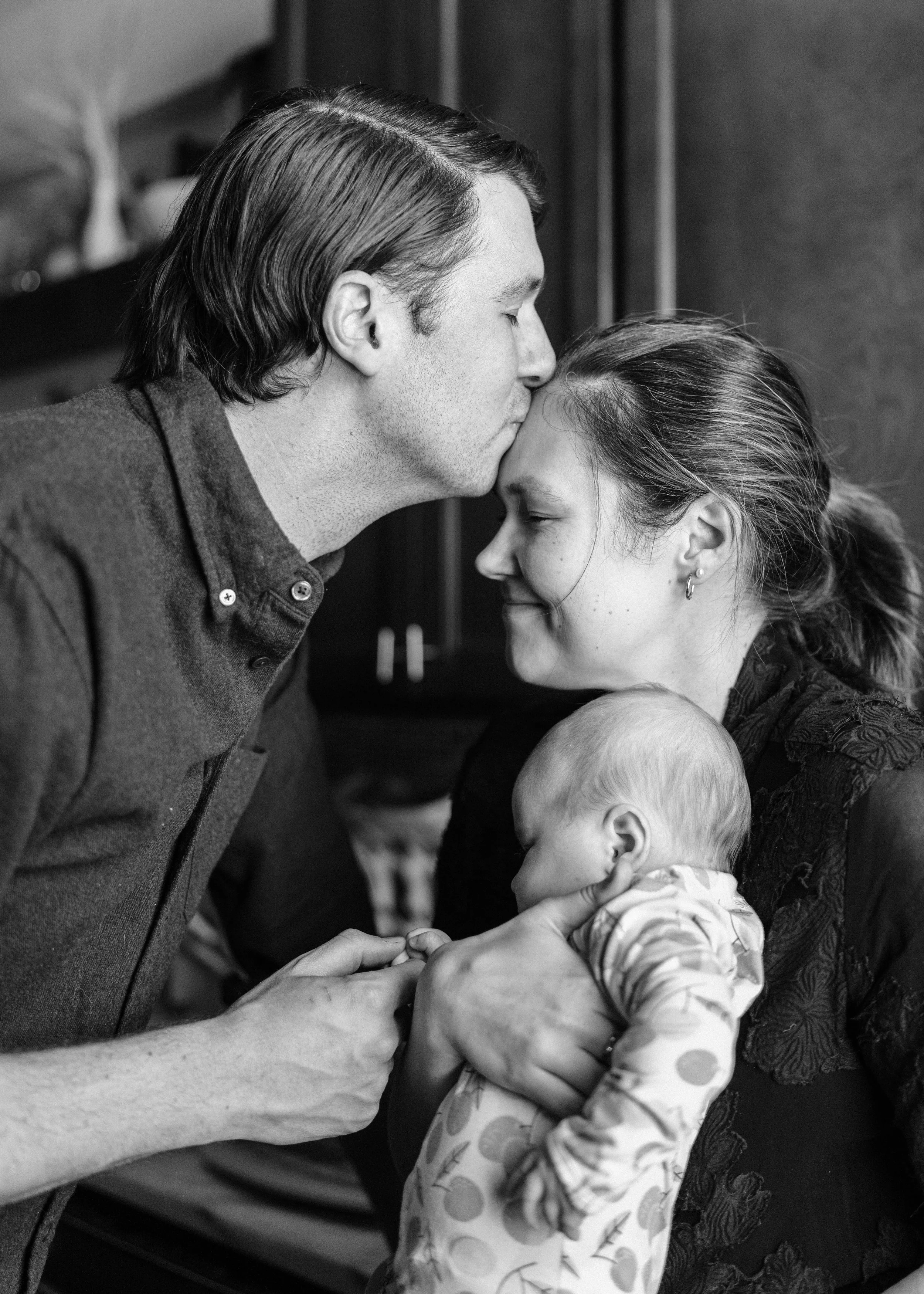 A black and white photo of a man sharing a gentle kiss on the forehead with a woman, who is holding a baby. The woman has her eyes closed with a content expression, and the baby is resting peacefully in her arms.