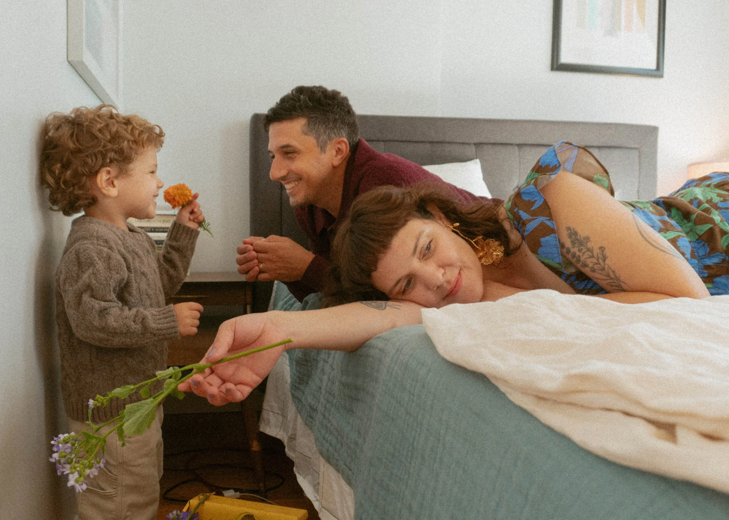 A young boy giving a flower to a man who is smiling. A woman is lying on the bed, holding a flower, with a happy expression. The scene shows a cozy bedroom setting with a bedside table and artwork on the wall.