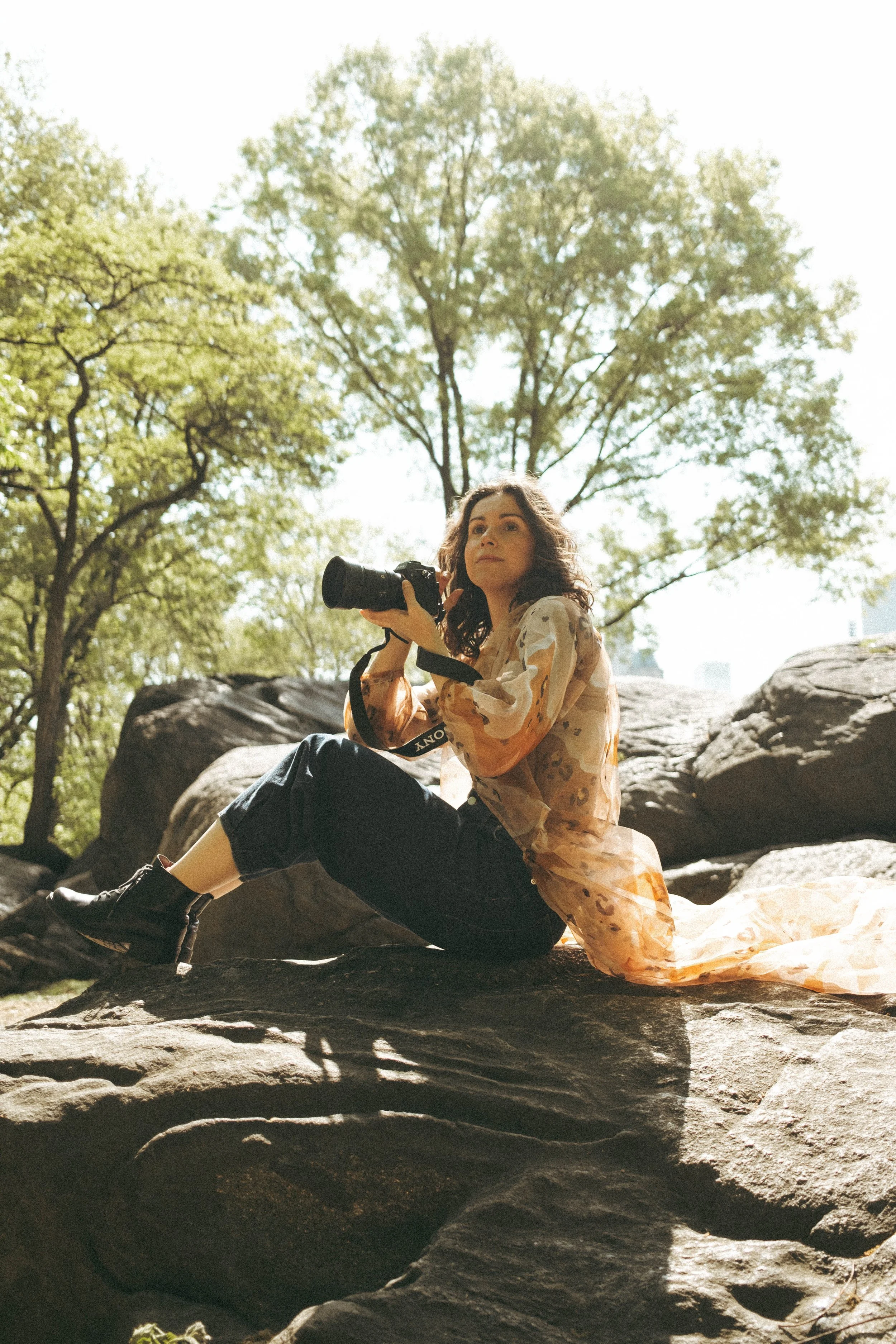 A woman with curly hair sitting on a large rock, holding a camera, outdoors in a park with green trees and rocks around her.