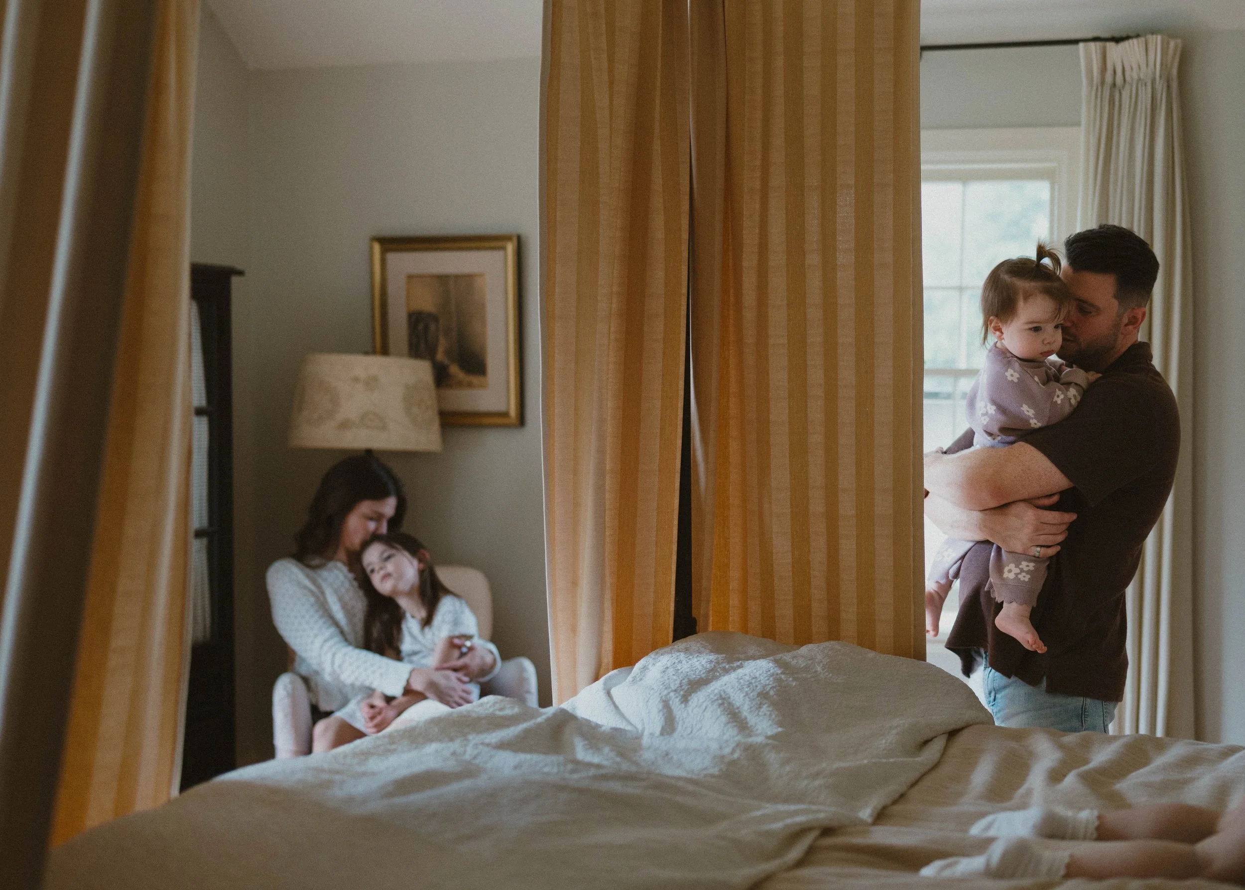 A man holding a young girl in pajamas, hugs her near a window with beige curtains, while a woman and two children sit on a bed in the background of a cozy bedroom.
