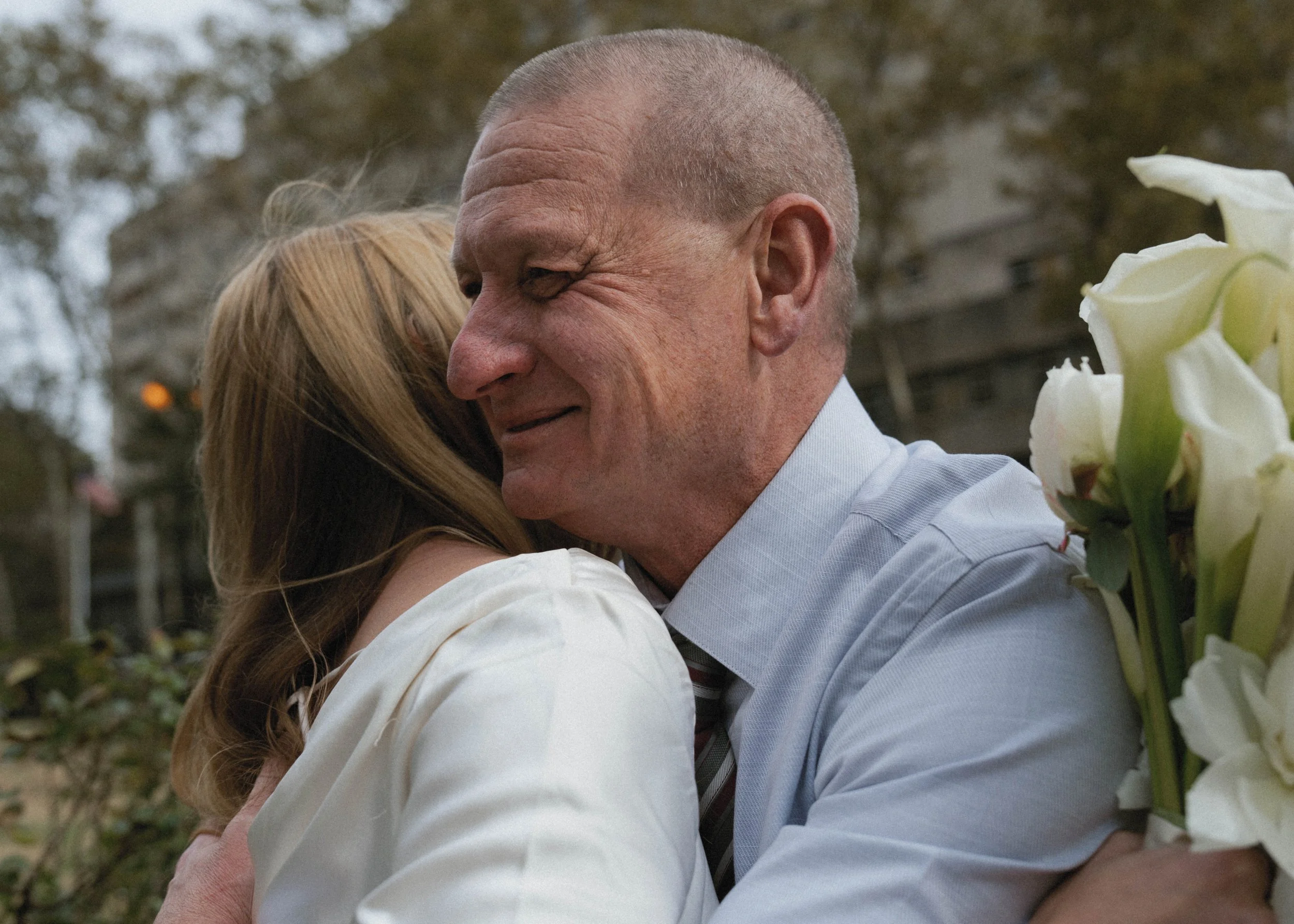 A man and woman embrace outdoors, the man smiling with his eyes closed and holding a bouquet of white flowers.